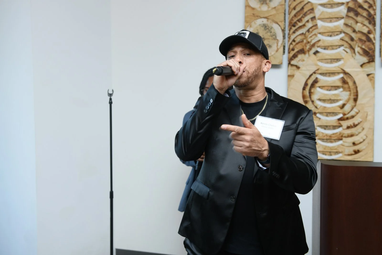 Man in black jacket and cap singing into microphone at an indoor event with abstract wooden wall art in background.