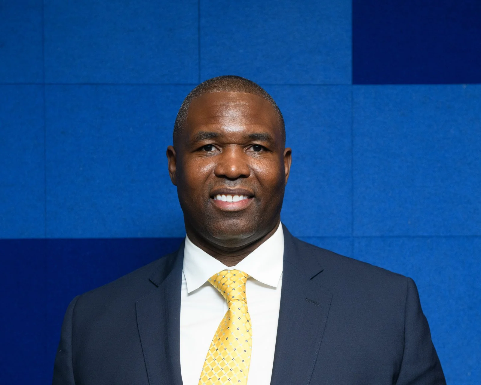 A man in a dark suit and yellow tie smiling in front of a blue wall.