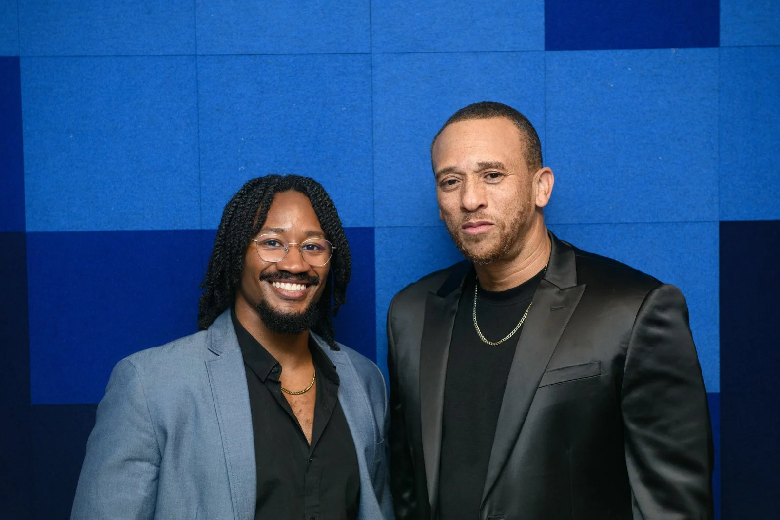 Two men standing in front of a blue wall, both dressed in black tuxedos with black shirts, one with glasses and the other with a chain necklace, smiling and looking at the camera.