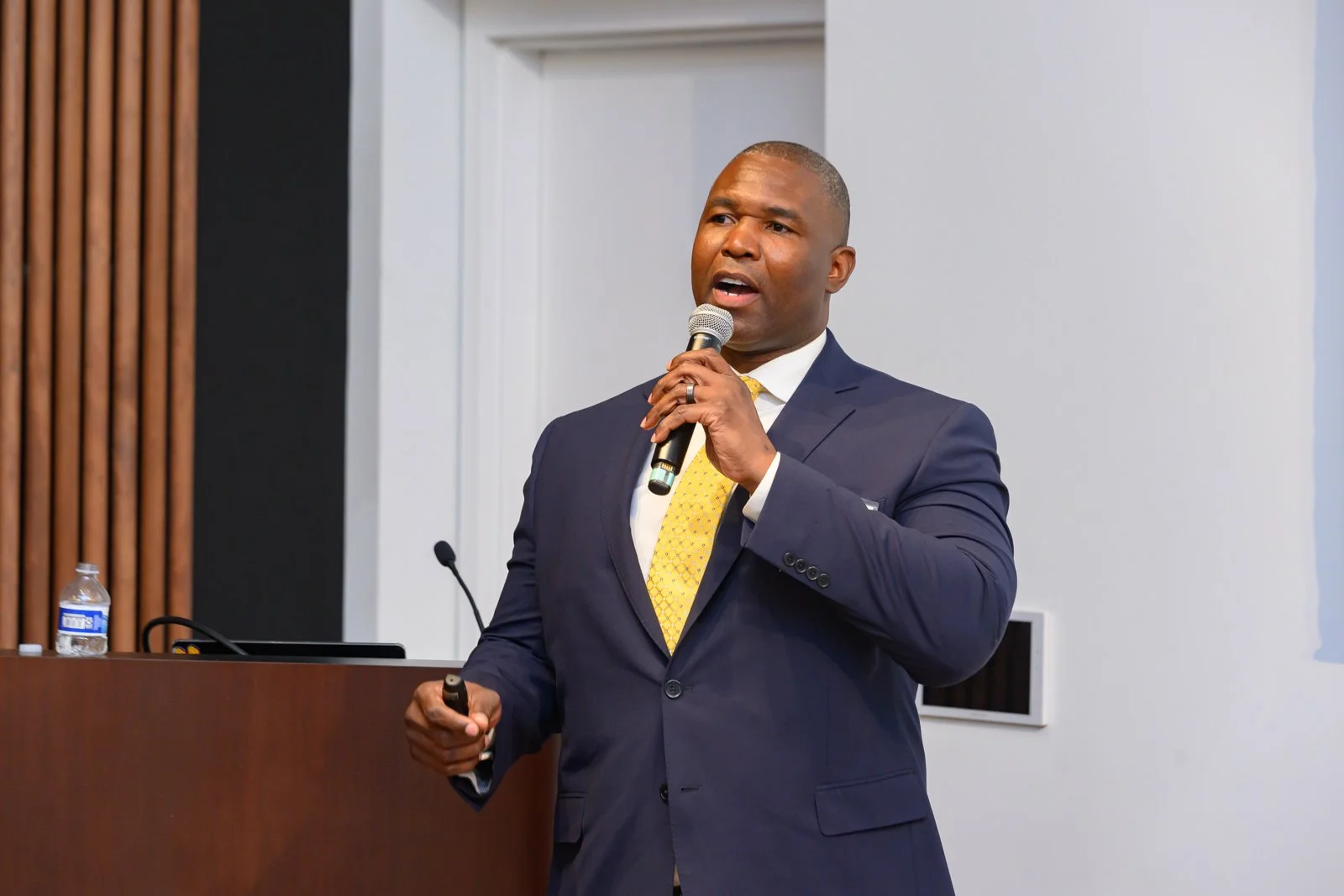 A man in a navy suit and yellow tie giving a presentation, holding a microphone in one hand and a remote in the other, with a water bottle on a table behind him.