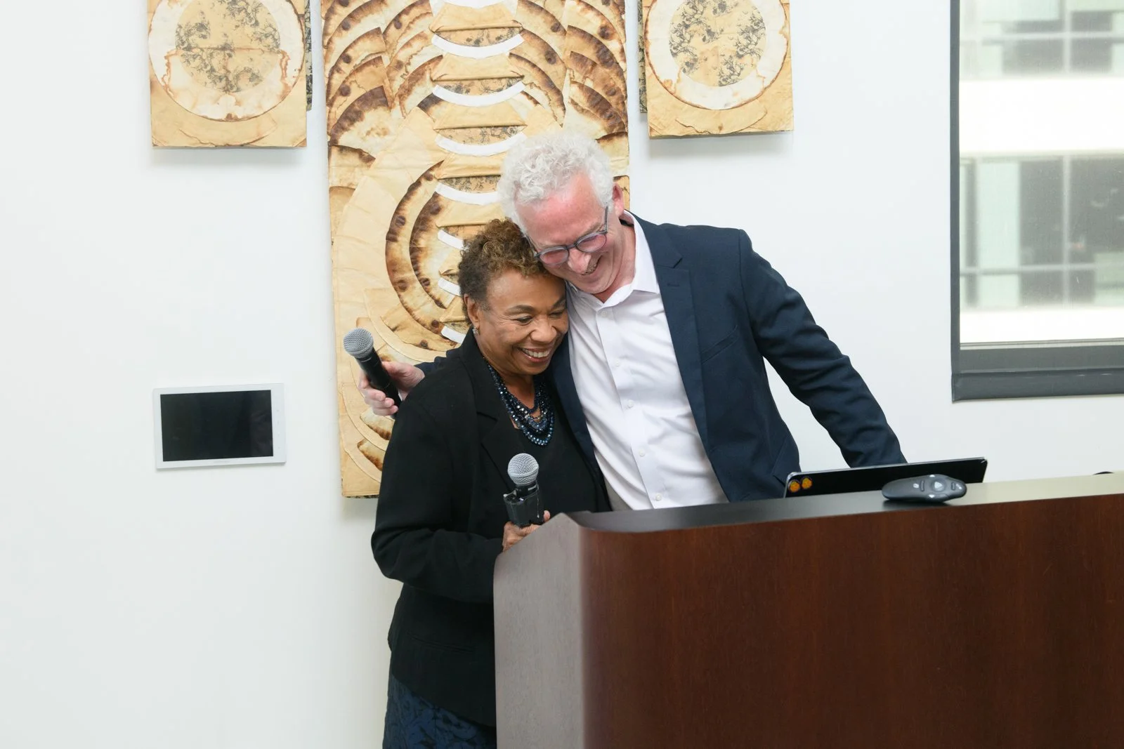 Two smiling women, one African American and one Caucasian, share a joyful moment while holding microphones at a podium during an event in a modern office or conference room with abstract artwork on the wall behind them. They are leaning close, lookin