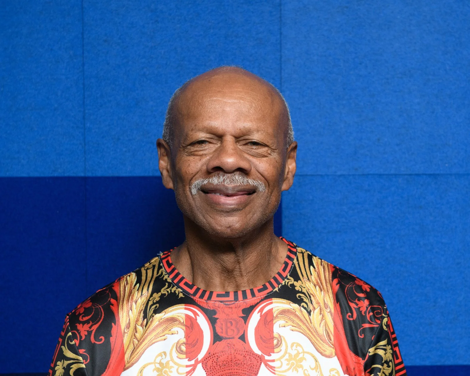 A smiling elderly man with a bald head and white mustache, wearing a patterned shirt with black, red, and gold designs, standing against a blue textured background.