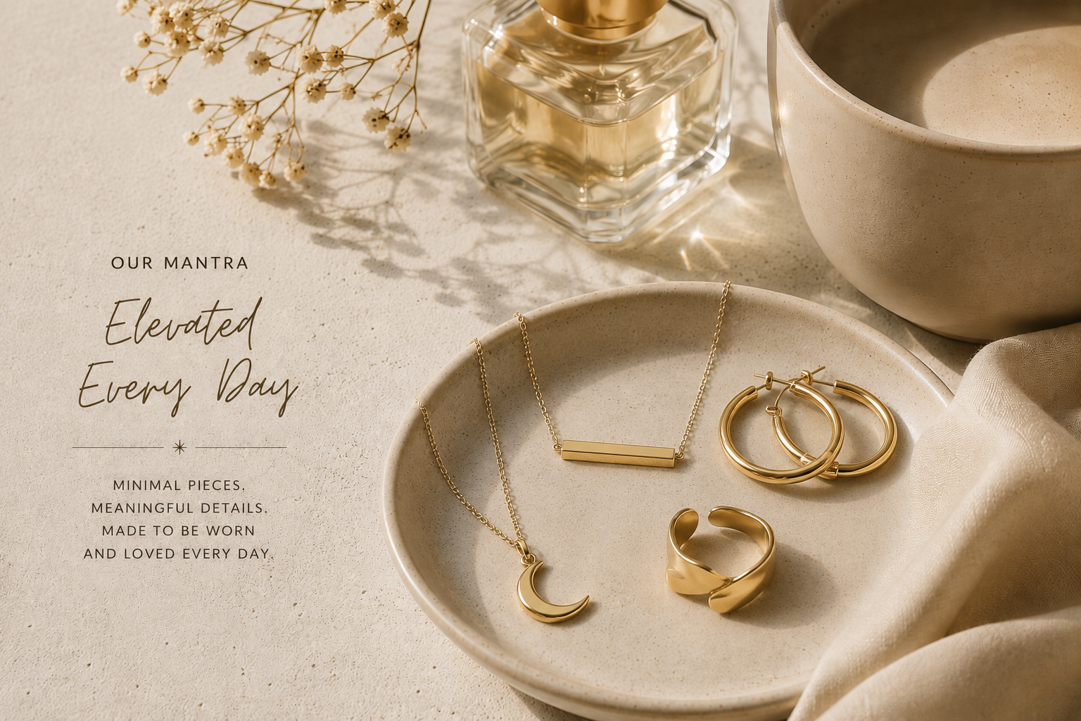 A beige ceramic dish holding gold jewelry, including a crescent moon necklace, a horizontal bar necklace, and two hoop earrings, with a beige ceramic mug, a glass perfume bottle, a sprig of baby's breath flowers, and a beige fabric on a textured surface.