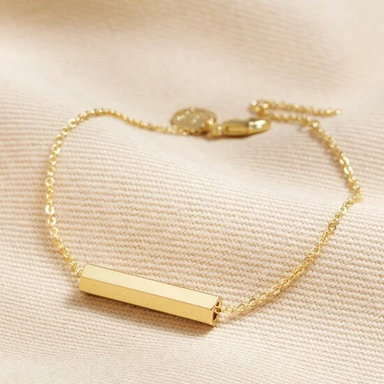 Gold bracelet with a rectangular bar on a soft, light-colored fabric background.