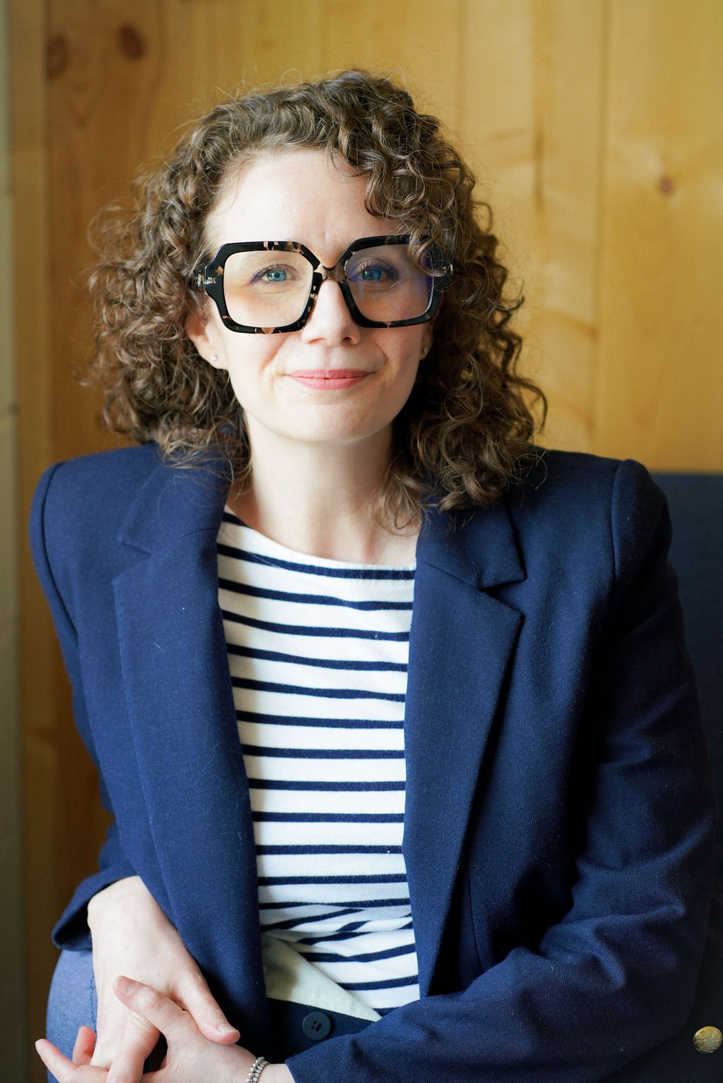 Meet Jessie - a woman with curly hair wearing large black glasses, a navy blazer, and a striped shirt, sitting in front of a wooden wall.