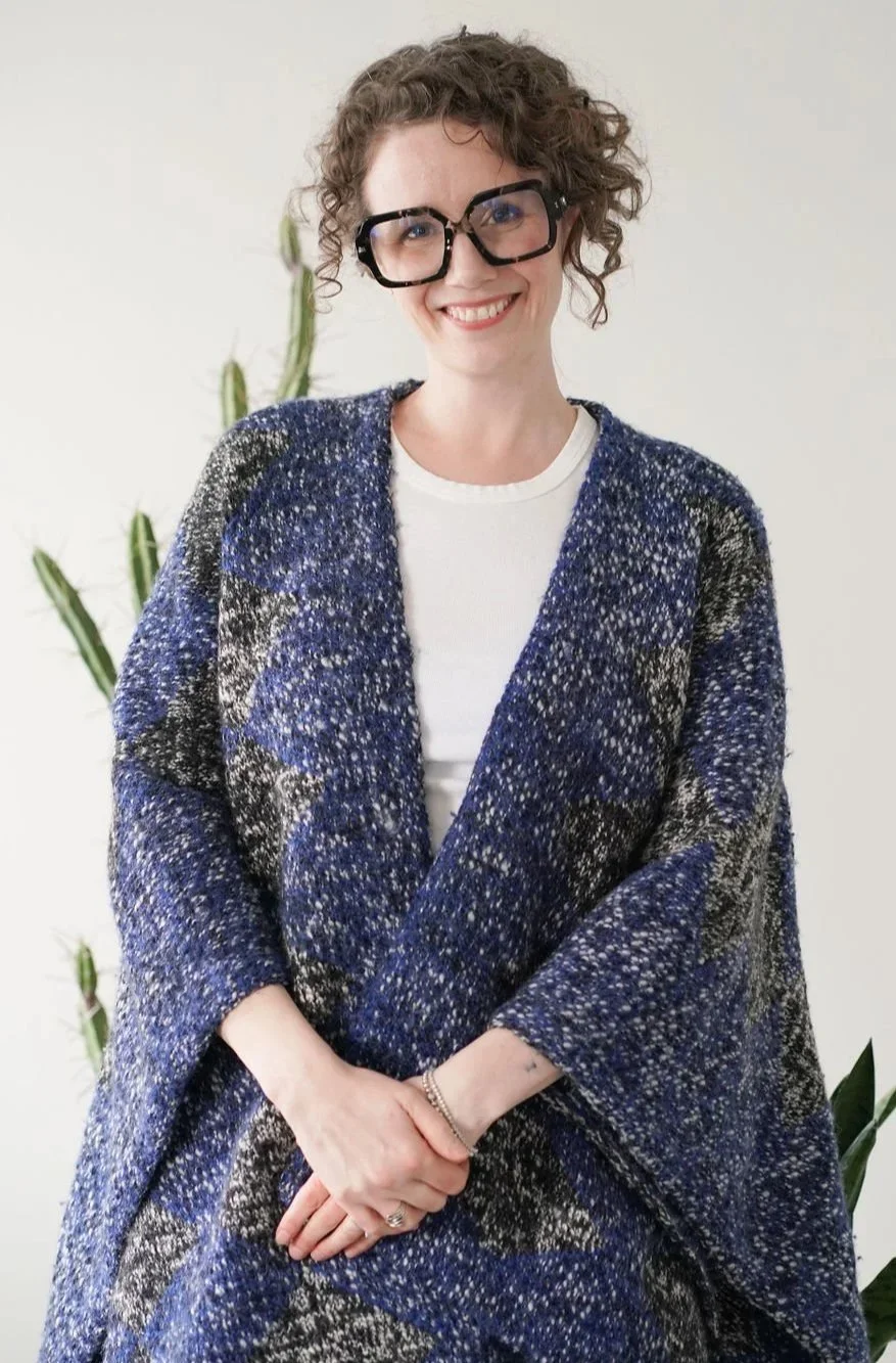 A woman with short curly hair and large black glasses smiling at the camera. She is wearing a white top and a blue, black, and white patterned shawl. There is a tall green cactus in the background and a white wall.
