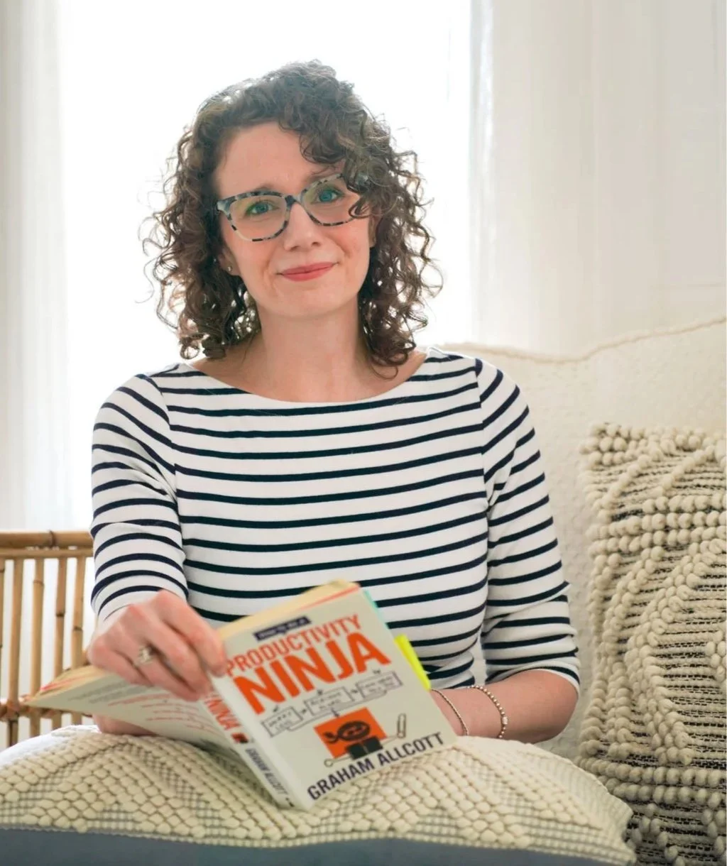 Jessiet sits on a woven blanket, holding a book titled 'Productivity Ninja' by Graham Allcott, inside a bright room with curtains and textured pillows.
