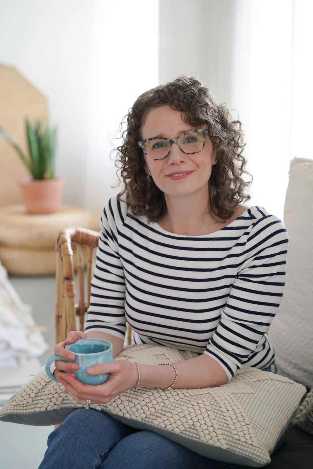 Jessie sitting on a couch, holding a blue mug, smiling at the camera in a cozy, well-lit room with houseplants and cushions.