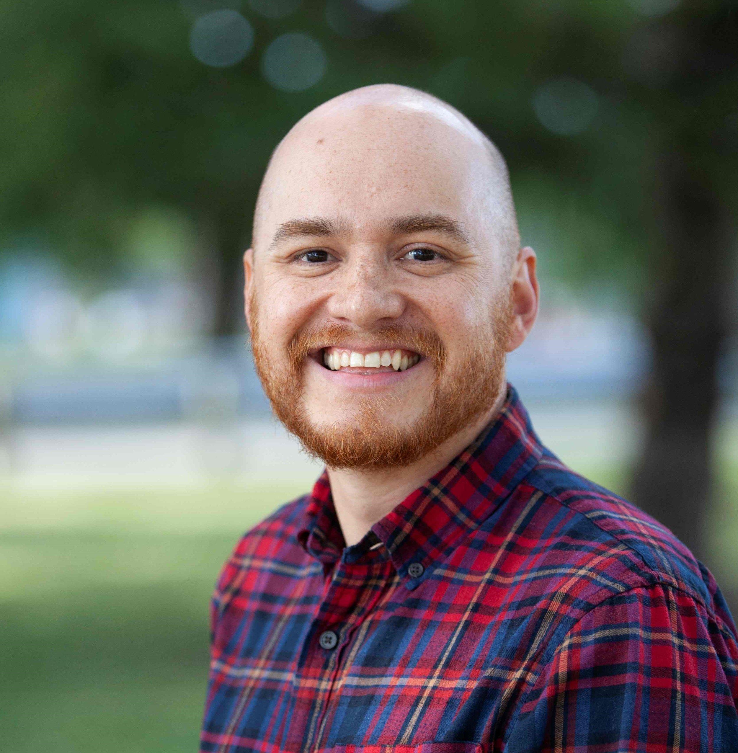 A smiling man with a shaved head, red hair, and a red beard, wearing a red and blue plaid shirt, outdoors with blurred green background.