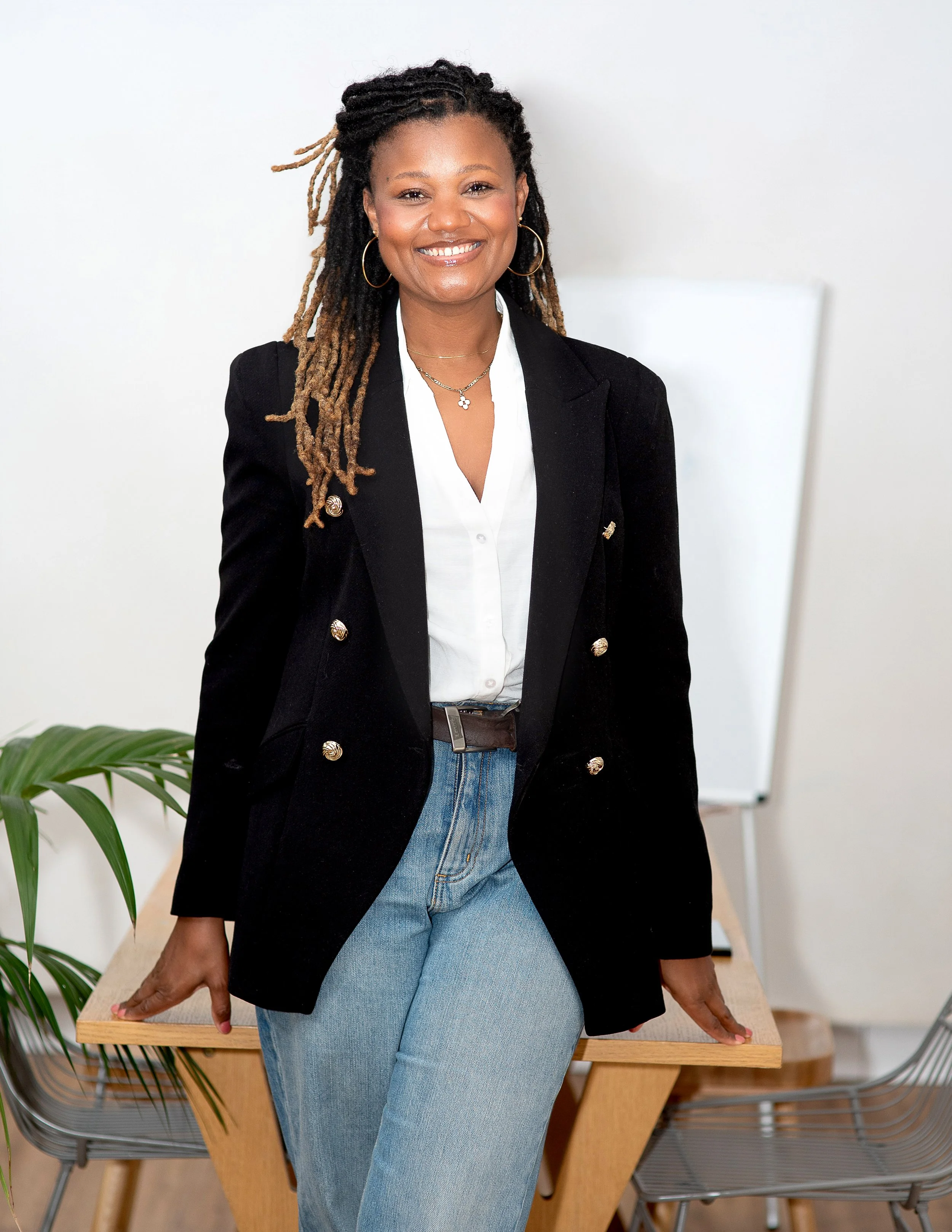 HR professional and executive coach Natallie Ateere wearing a black blazer, smiling while leaning on a wooden table in a modern office space.