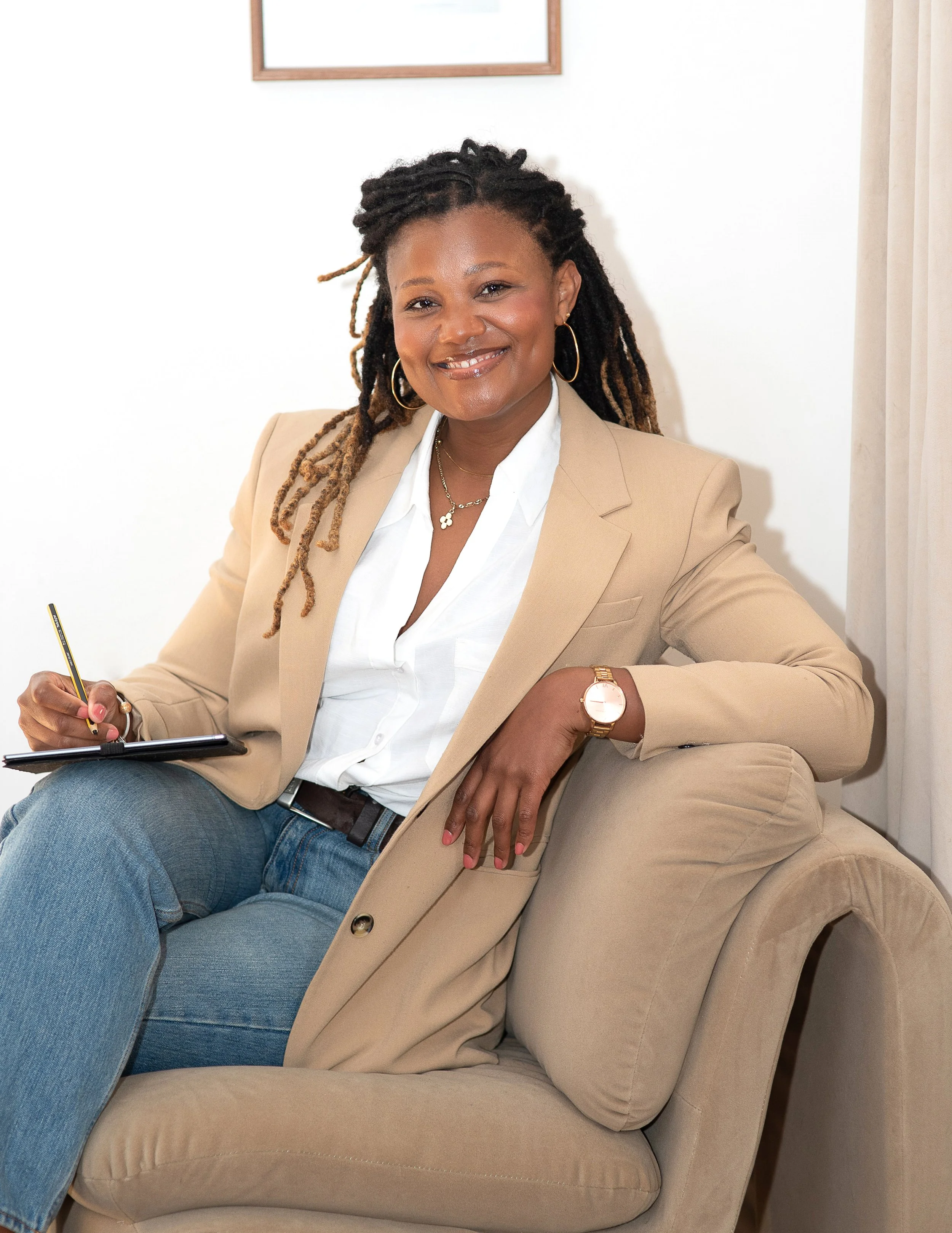 Executive and leadership coach Natallie Ateere smiling, wearing a beige blazer and white shirt, sitting on a beige couch with a pen and a notepad.