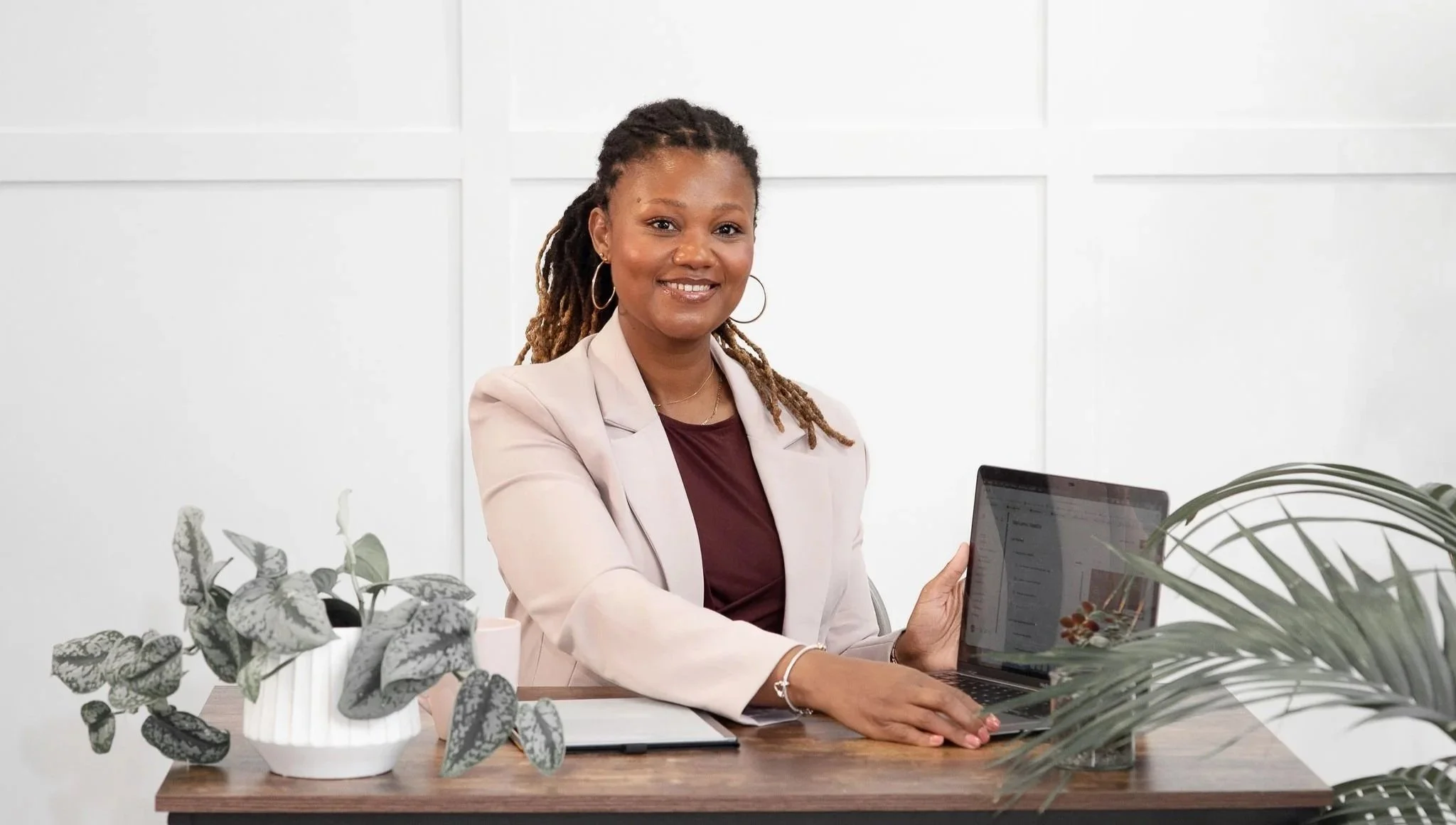 Executive coach Natallie Ateere with dreadlocks and hoop earrings smiling while sitting at a desk with a laptop, surrounded by houseplants in a bright office.