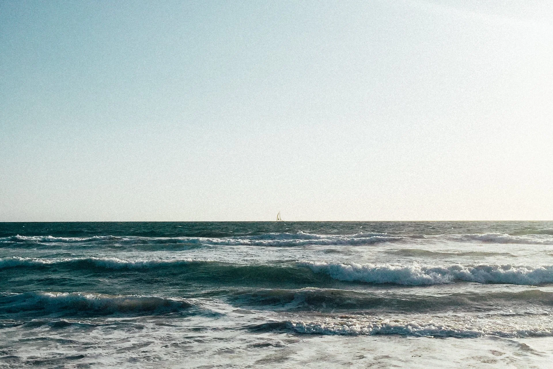 Ocean waves with a sailboat on the horizon under a clear sky.