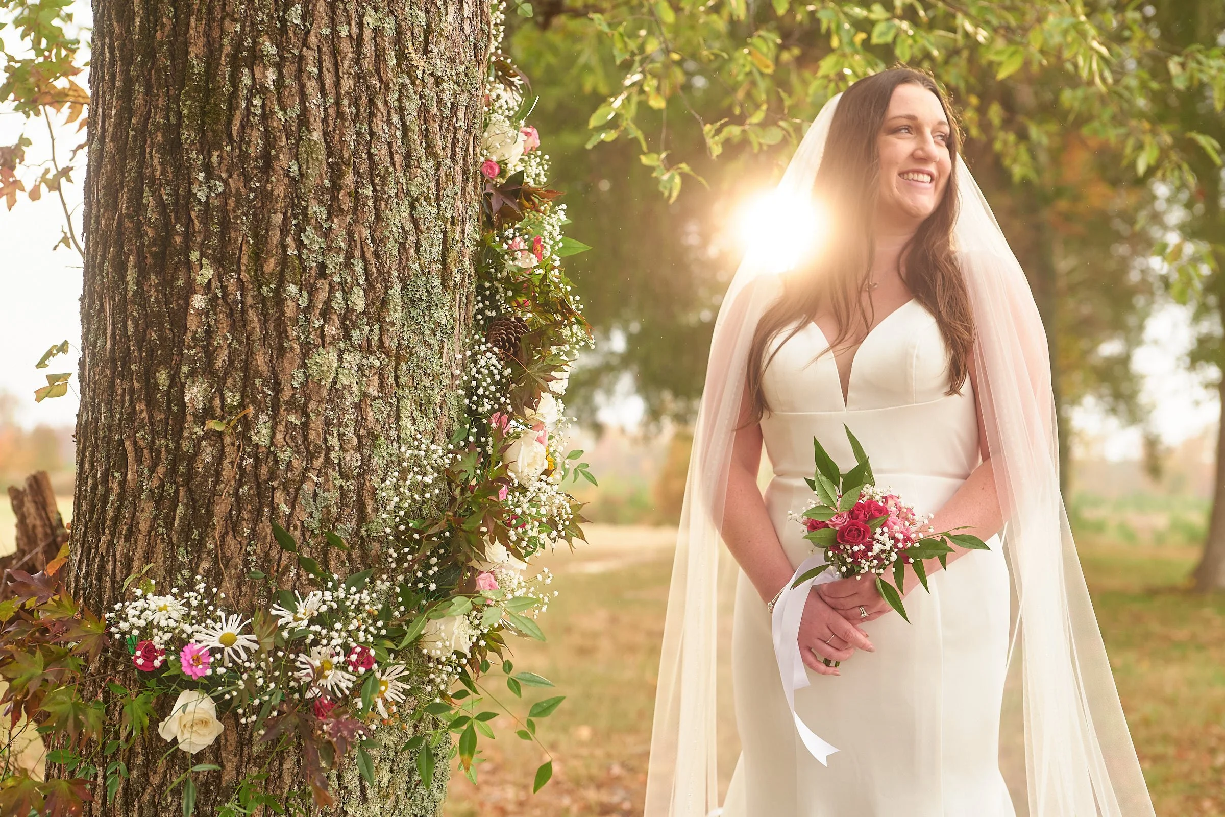 A bride standing outdoors near a tree decorated with flowers, smiling and holding a bouquet, with sunlight filtering through the trees.