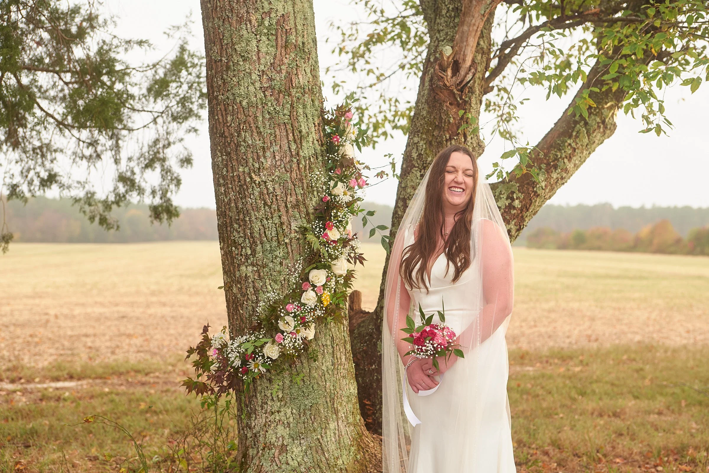 A woman in a wedding dress with a veil, smiling with her eyes closed, standing outdoors near a tree decorated with flowers, in a field with trees in the background.