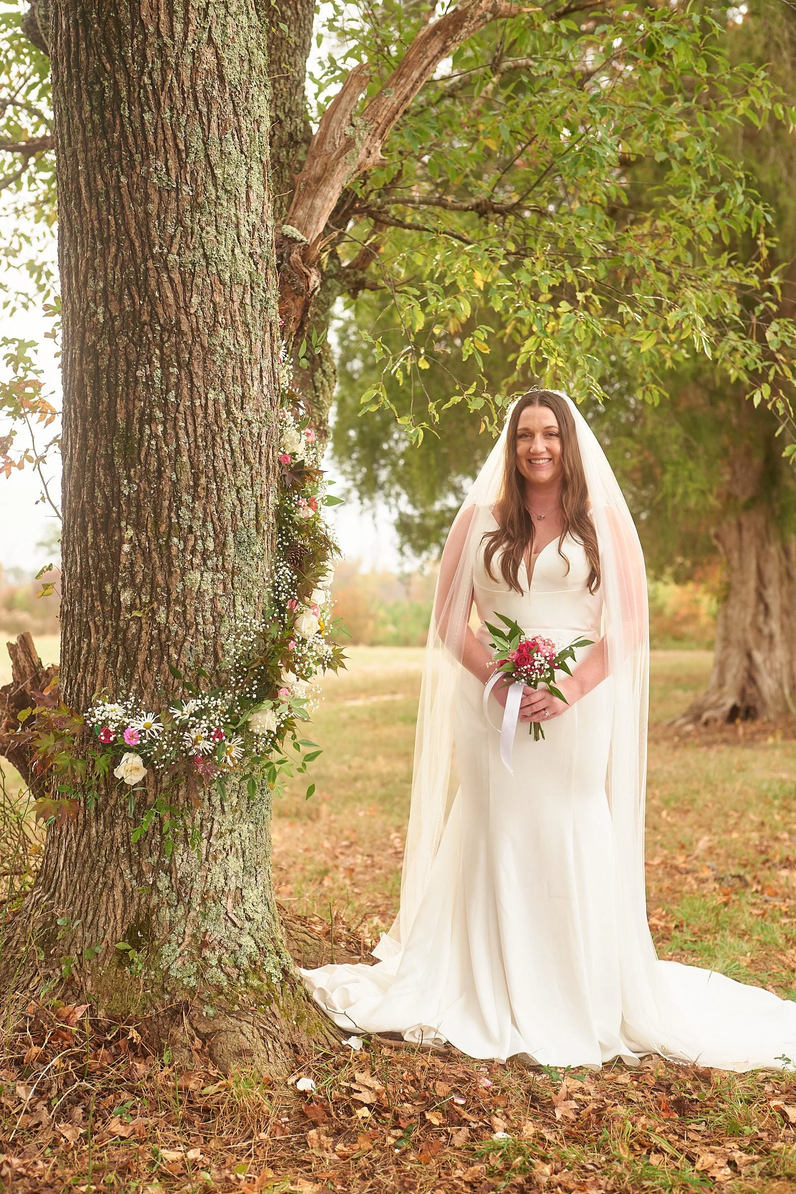 A bride in a white wedding gown standing outdoors near a large tree with a floral decoration on the trunk, holding a bouquet, and smiling.