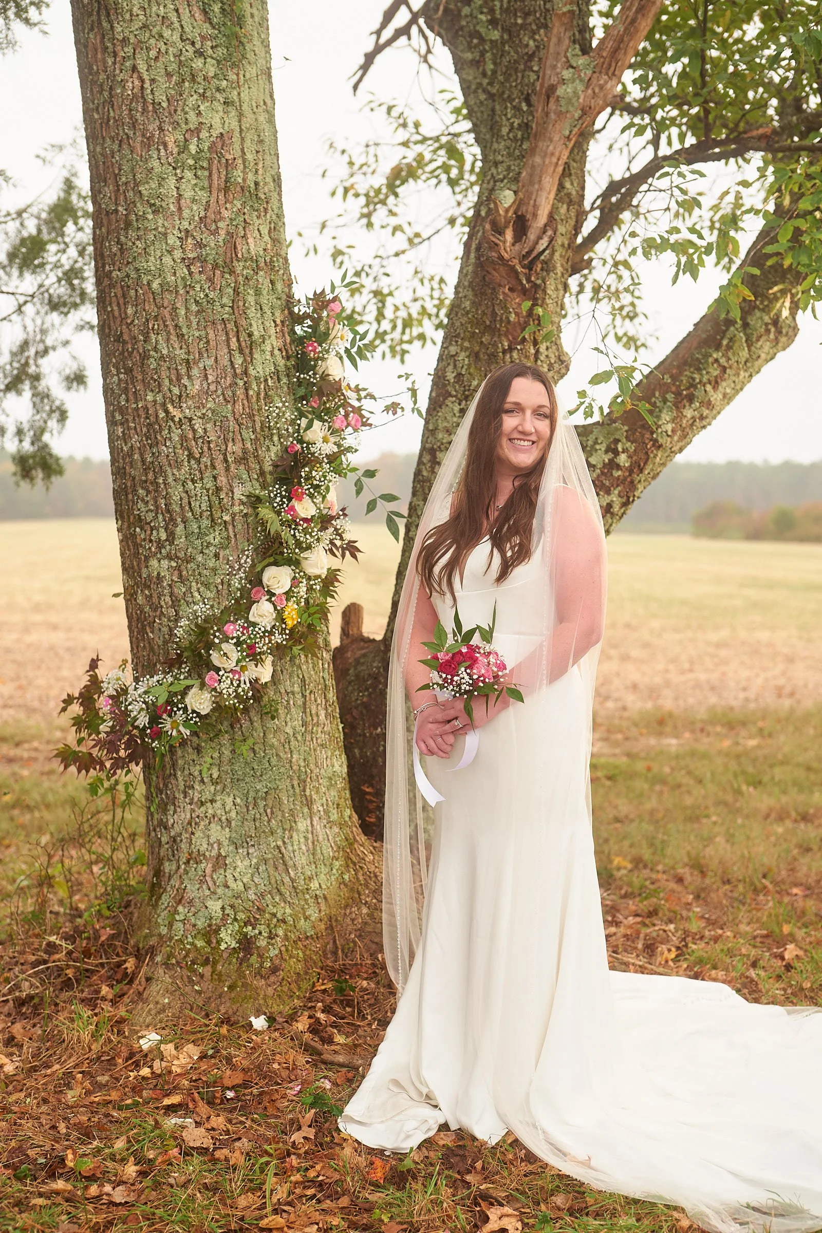 A smiling bride in a white wedding dress holding a bouquet stands outside near a tree decorated with flowers in an outdoor setting during the daytime.