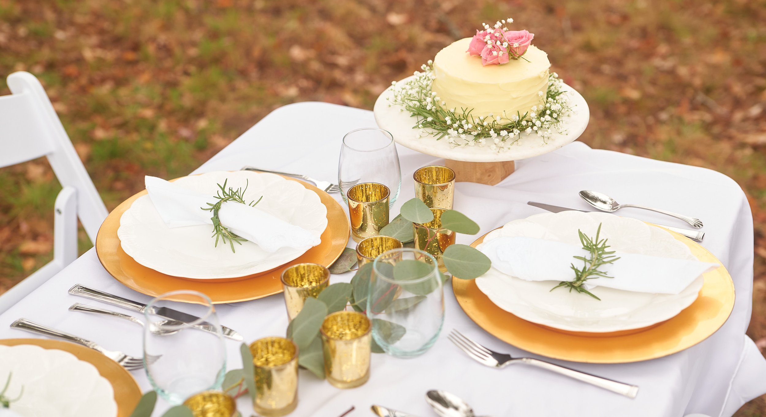 A table set for an outdoor celebration with white plates, gold chargers, clear glasses, and golden candle holders. A white cake decorated with pink flowers is at the center, surrounded by greenery and small white flowers, on a rustic wooden cake stan