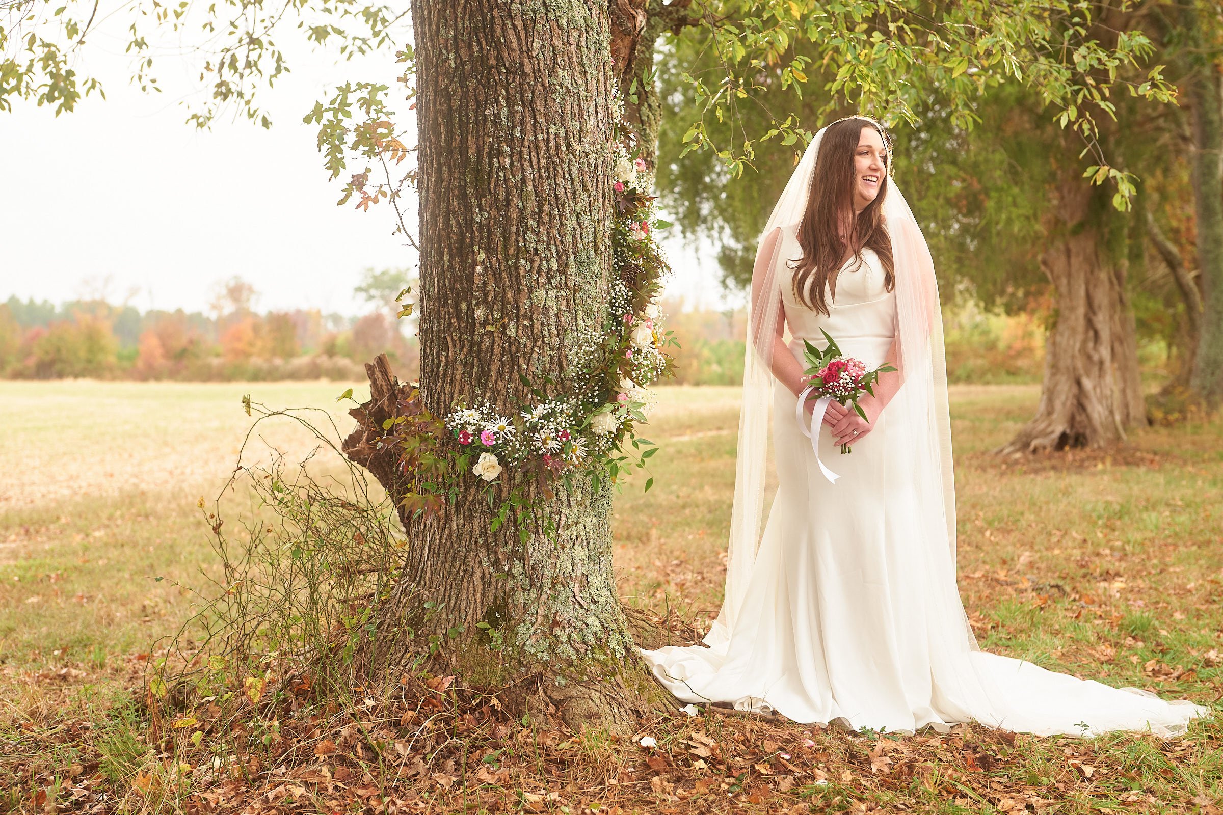 A bride in a white wedding dress and veil standing outdoors next to a large tree decorated with flowers and greenery in a natural setting with trees and open land in the background.