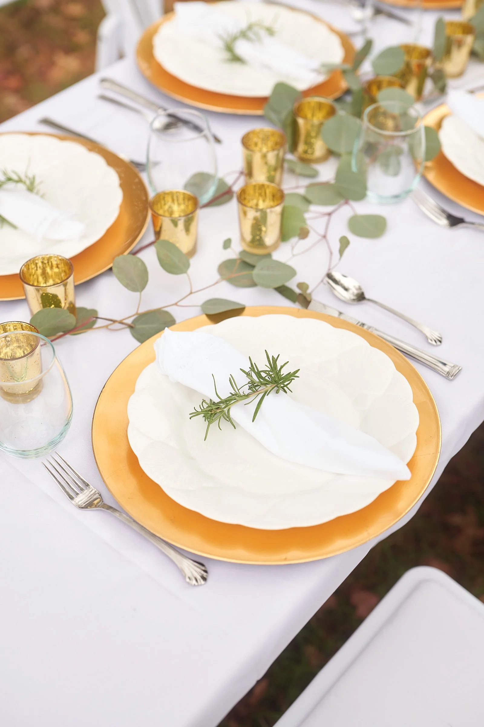 Elegant outdoor table setting with white plates, gold-rimmed plates, silverware, clear drinking glasses, gold candle holders, and eucalyptus leaves, with a white tablecloth and a sprig of rosemary as decoration.