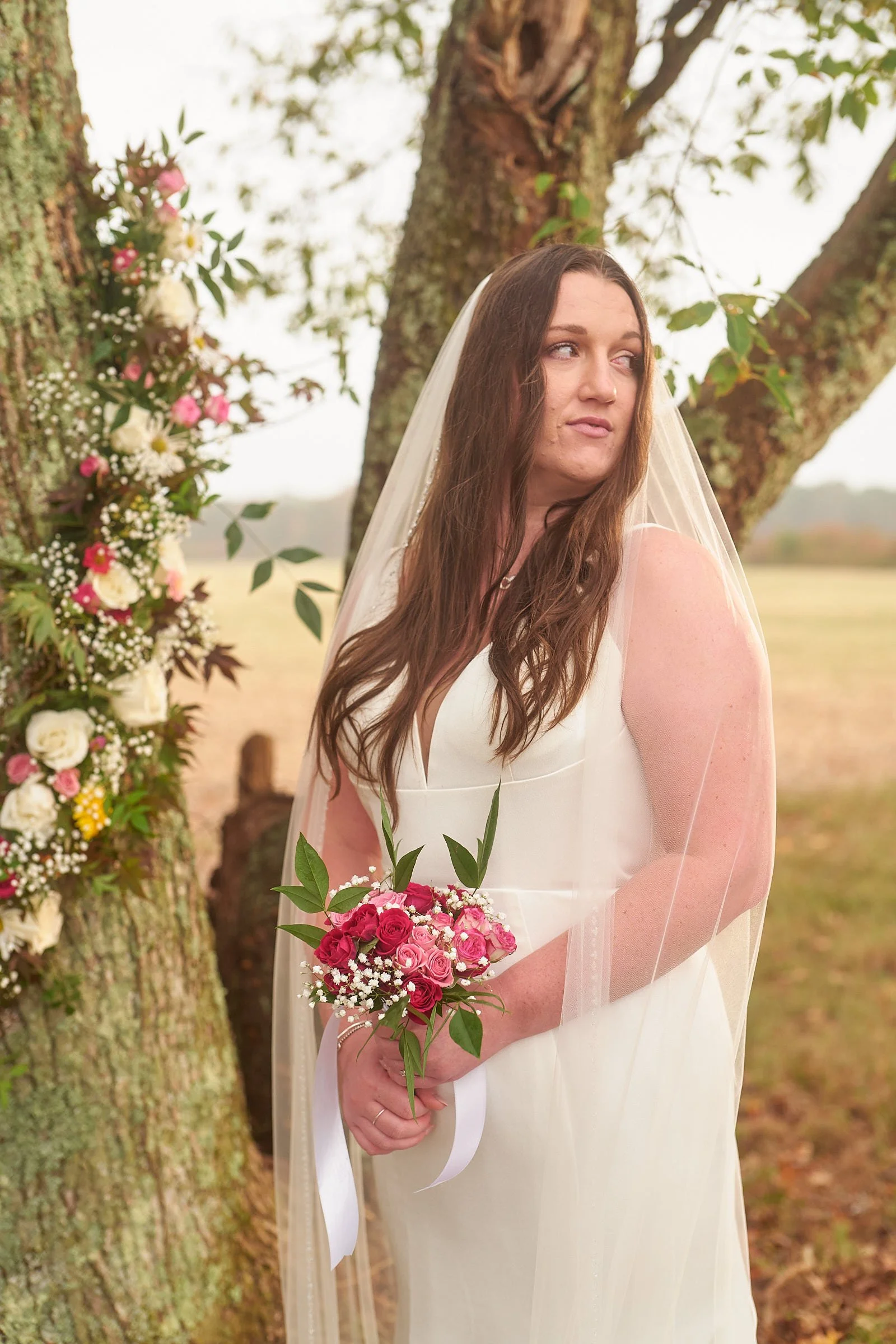 A woman in a white dress and veil holding a bouquet of pink and red roses with greenery, standing outdoors near a tree with decorated flowers, during daytime.