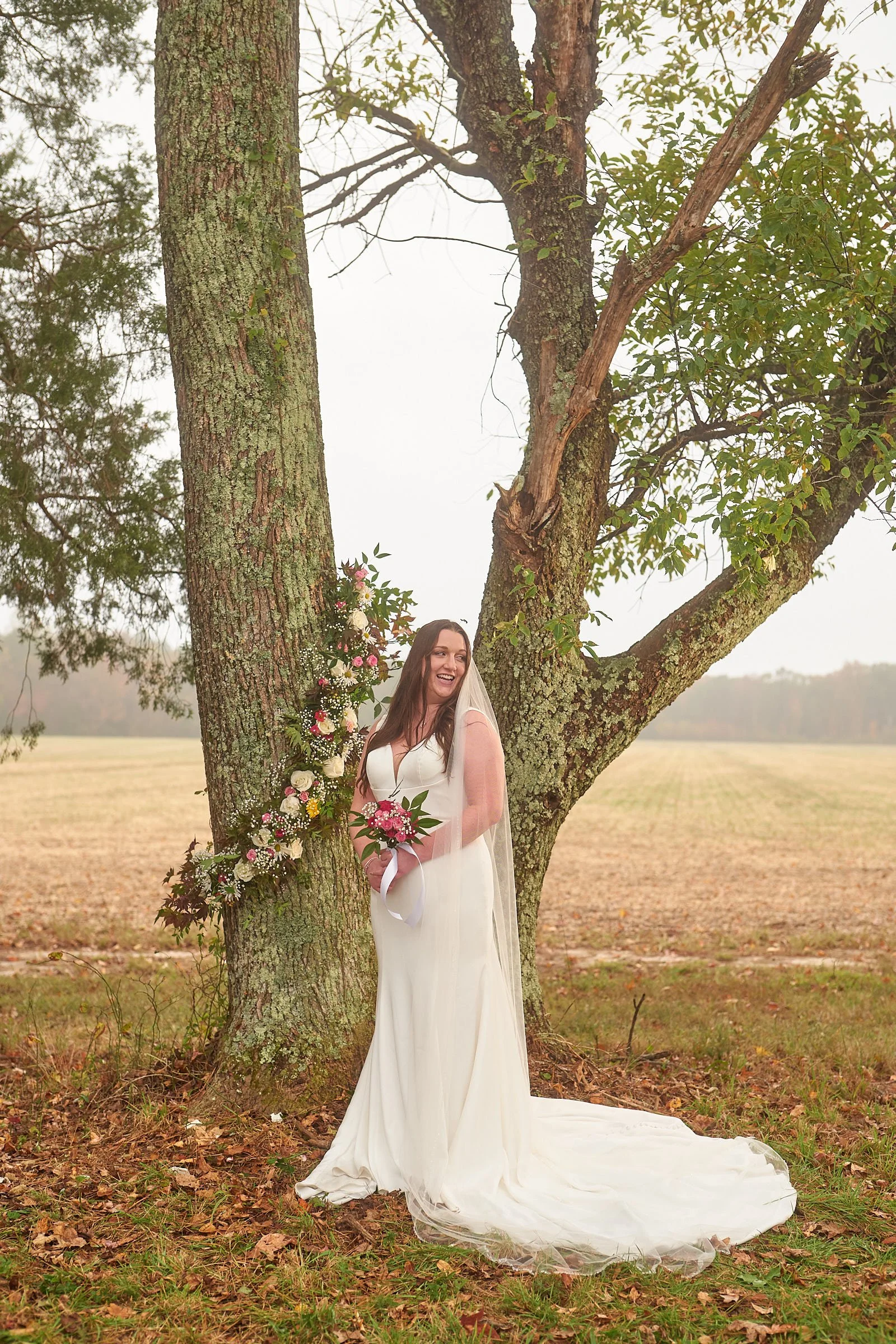 A woman in a white wedding dress standing outdoors near a tree with a flower arrangement attached to the tree trunk. She is holding a bouquet of pink and white flowers and smiling, with a natural open field in the background.