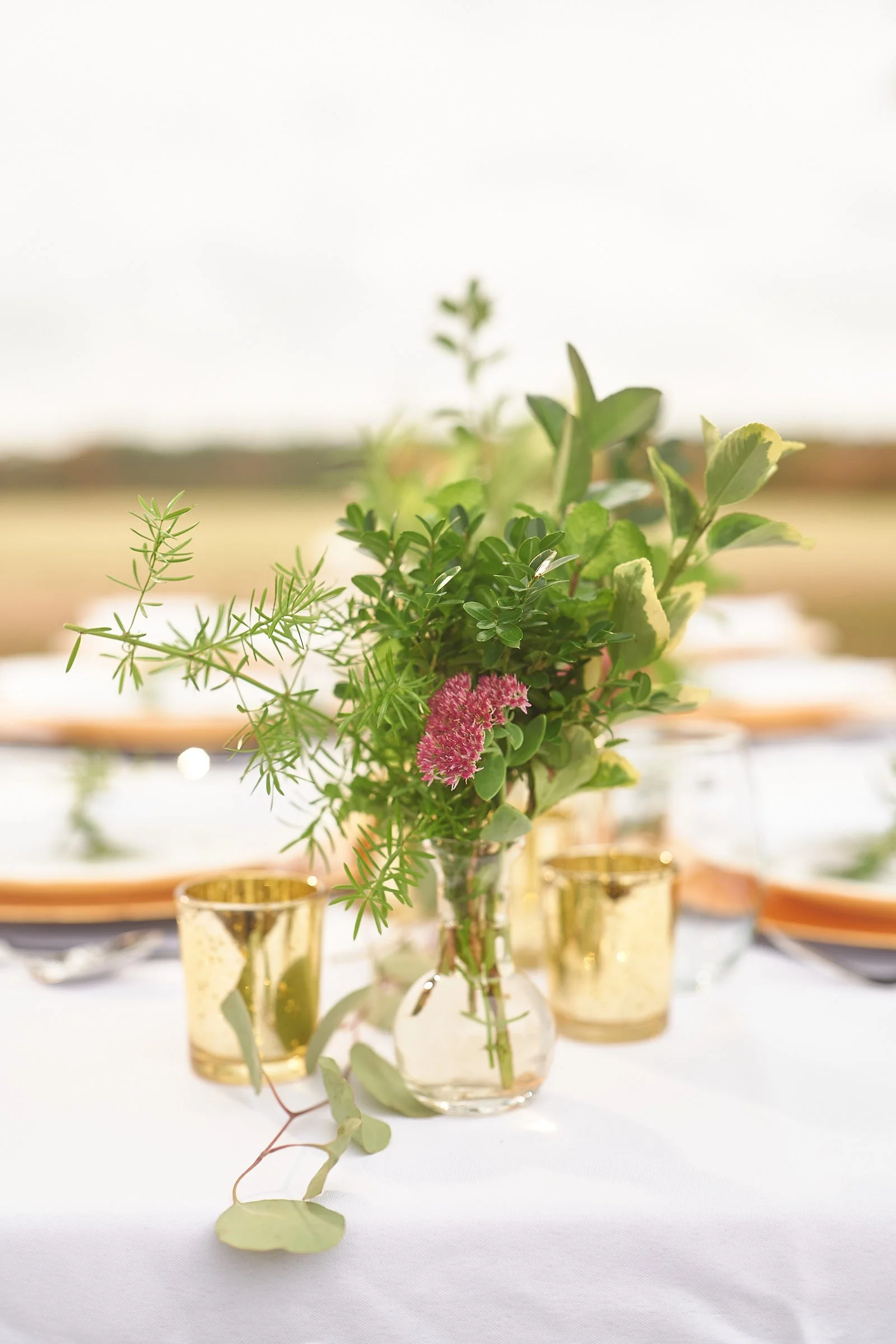 A floral centerpiece with green foliage and pink flowers arranged in glass vases on a white tablecloth, with golden cups around it.