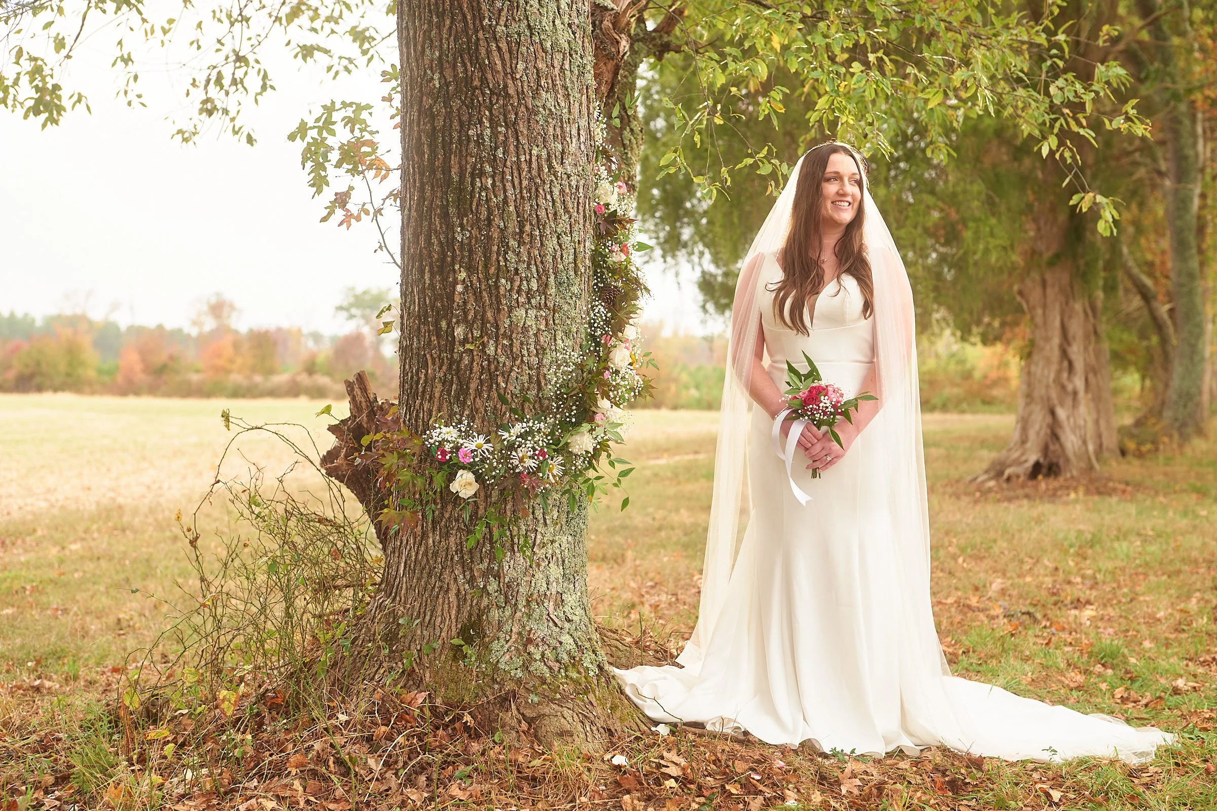 A woman dressed in a white wedding gown holding a bouquet, standing outdoors near a tree decorated with flowers in a natural setting with trees and open field in the background.