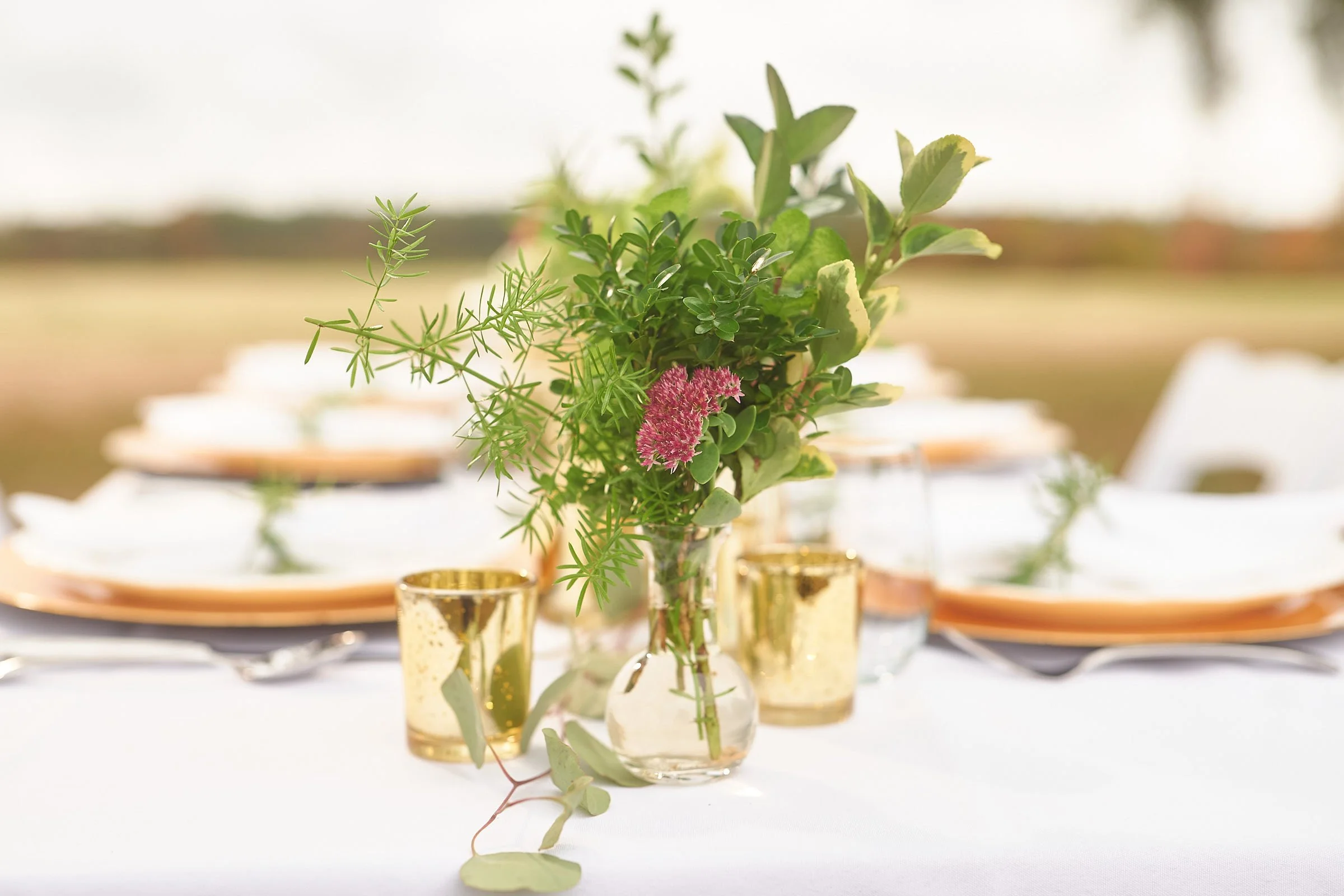 A floral centerpiece with green foliage and pink flowers in glass vases arranged on a white table, with gold accents and plates in the background.