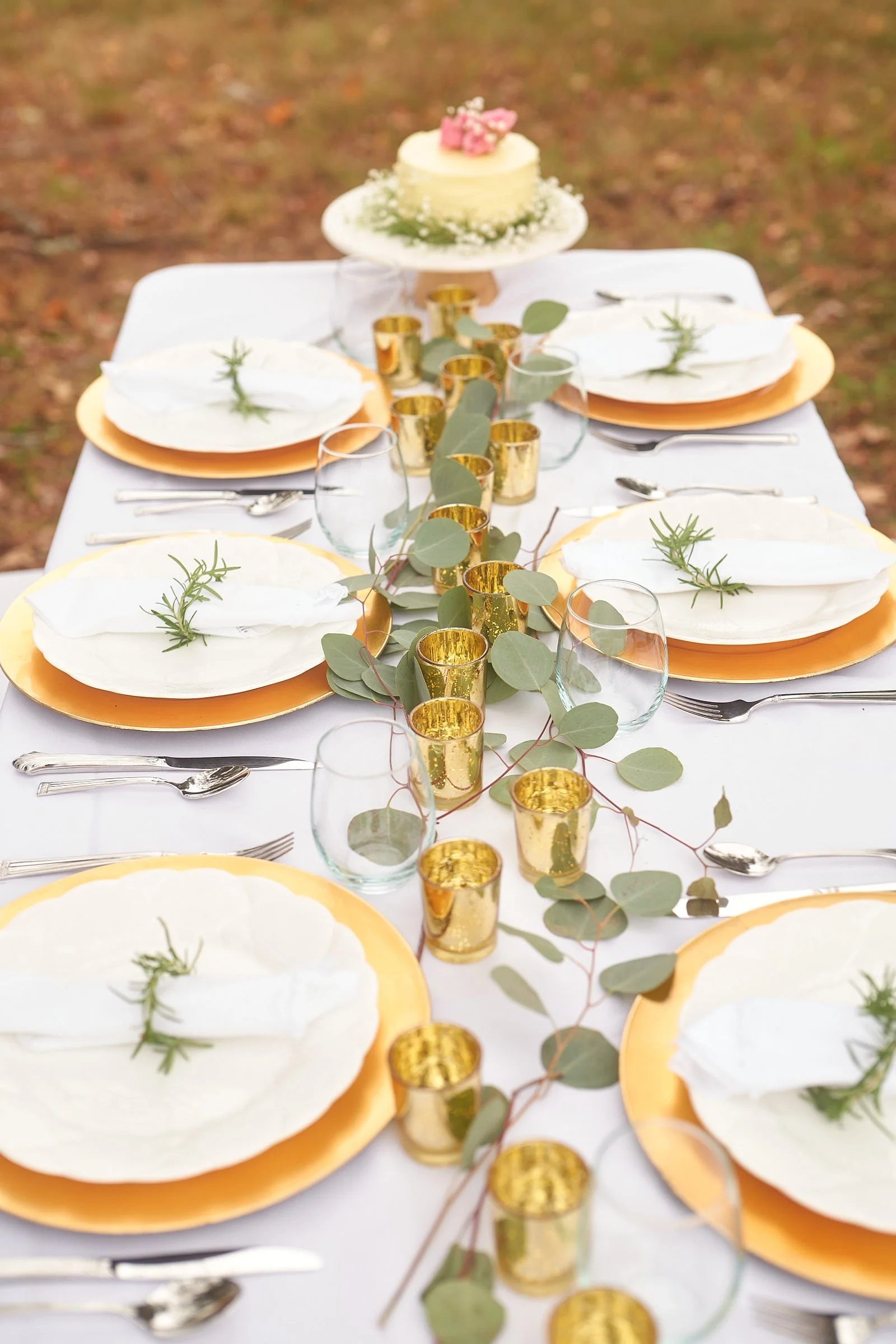 A decorated outdoor dining table set with white plates with gold rims, white napkins with small sprigs of rosemary, clear glasses, and gold candle holders, with a white cake topped with pink flowers in the background.