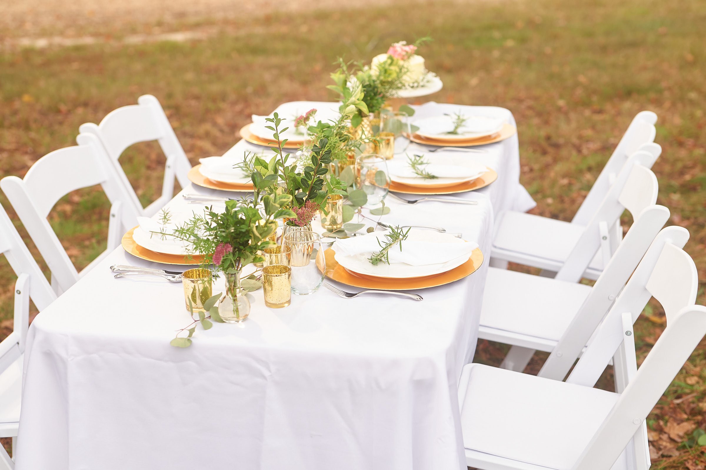 A outdoor wedding or event table setting with a white tablecloth, gold and glassware, white plates with green leaves, and centerpieces of flowers and greenery, surrounded by white chairs on grass.
