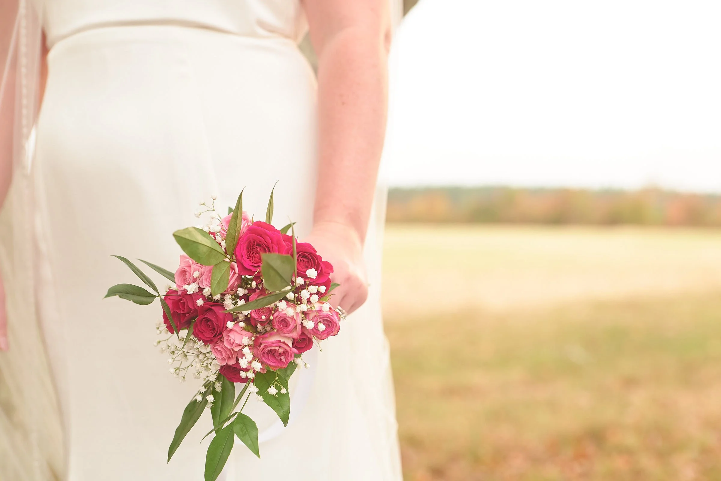 A person wearing a wedding dress holding a bouquet of pink and white roses with greenery, standing outdoors in a field with a blurred landscape in the background.