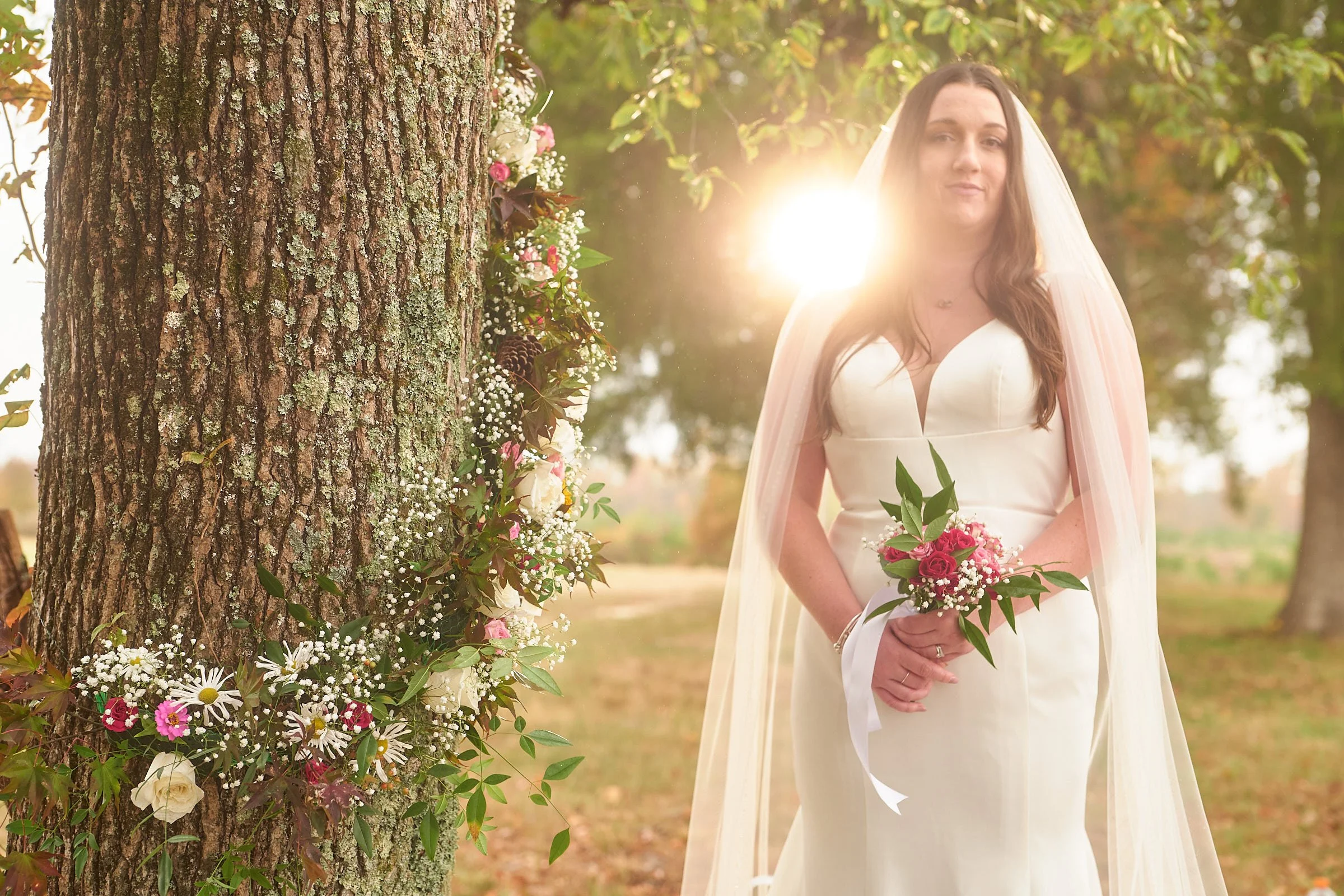 A woman dressed in a wedding gown holding a bouquet of flowers stands outdoors near a large tree decorated with pink and white flowers, with the sun shining in the background.
