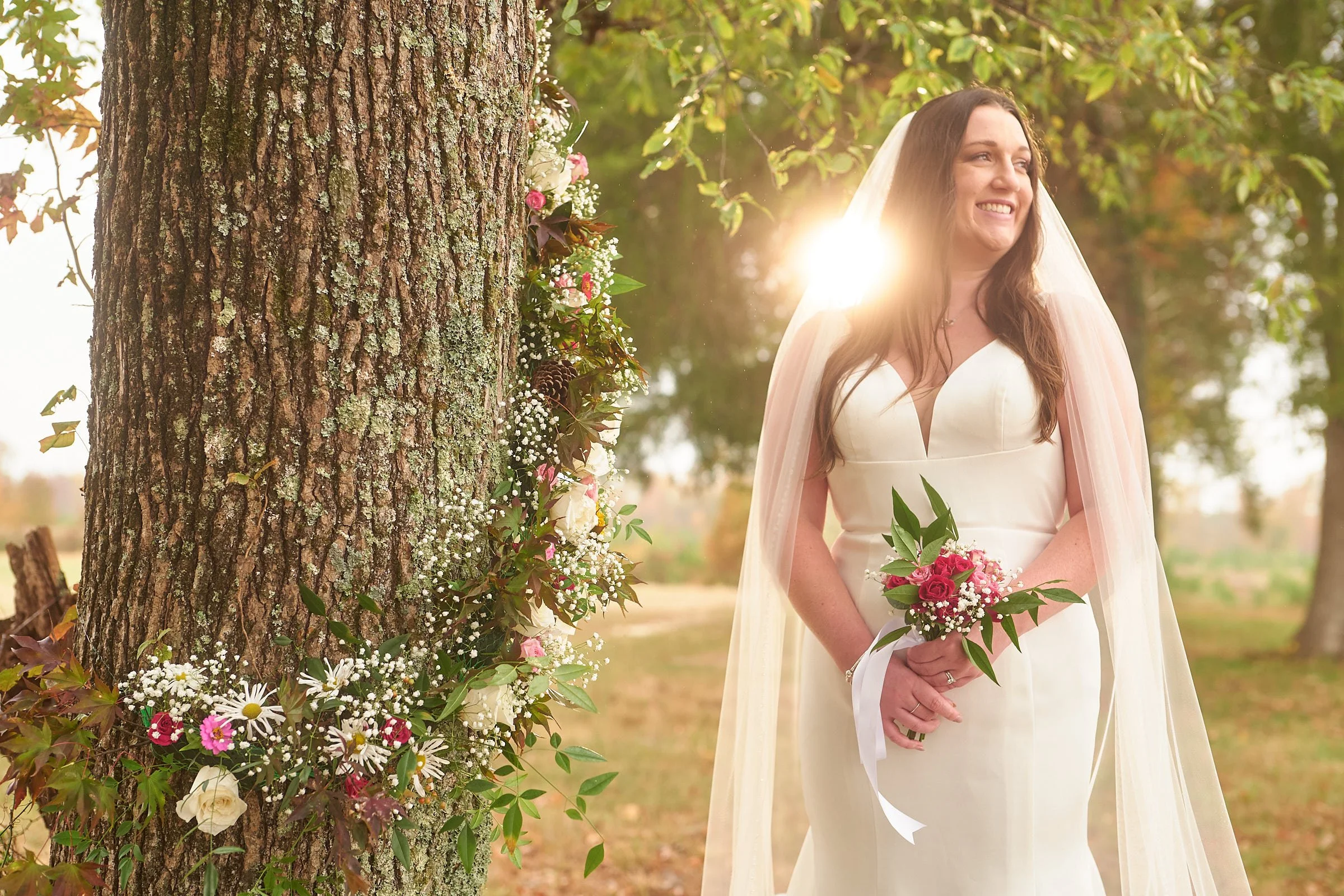 A smiling woman in a white dress and veil holding a bouquet of pink roses and other flowers, standing outdoors near a tree decorated with flowers and pinecones, sunlight filtering through the leaves.