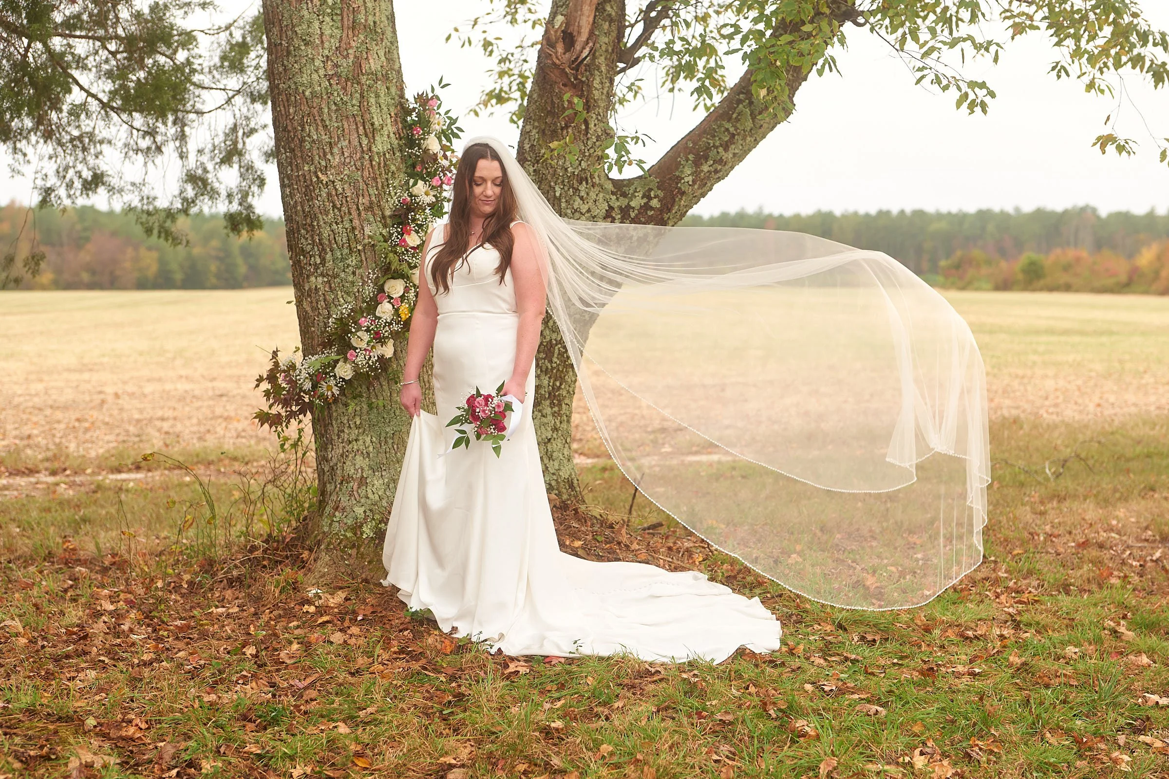 A bride in a white wedding dress holding a bouquet standing outdoors next to a large tree decorated with flowers, in a field with trees in the background.