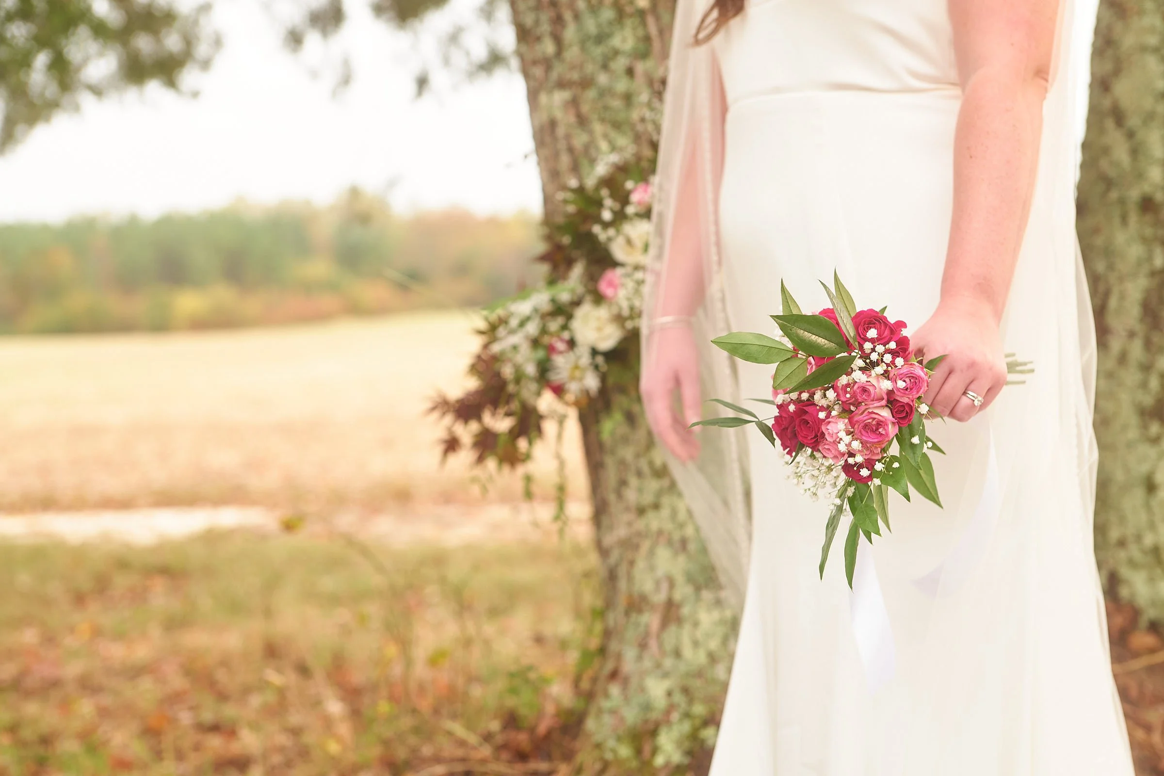 A bride dressed in a white wedding gown holding a bouquet of pink roses and white flowers outside near a tree.