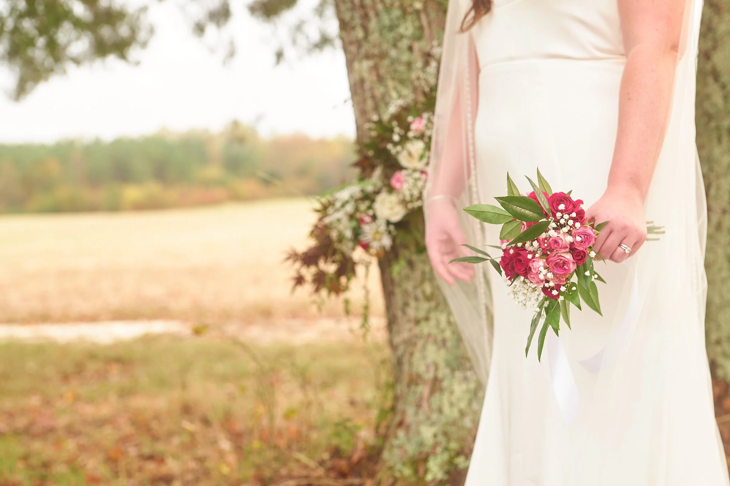 A woman in a white dress holding a pink and white flower bouquet, standing outdoors near a tree.