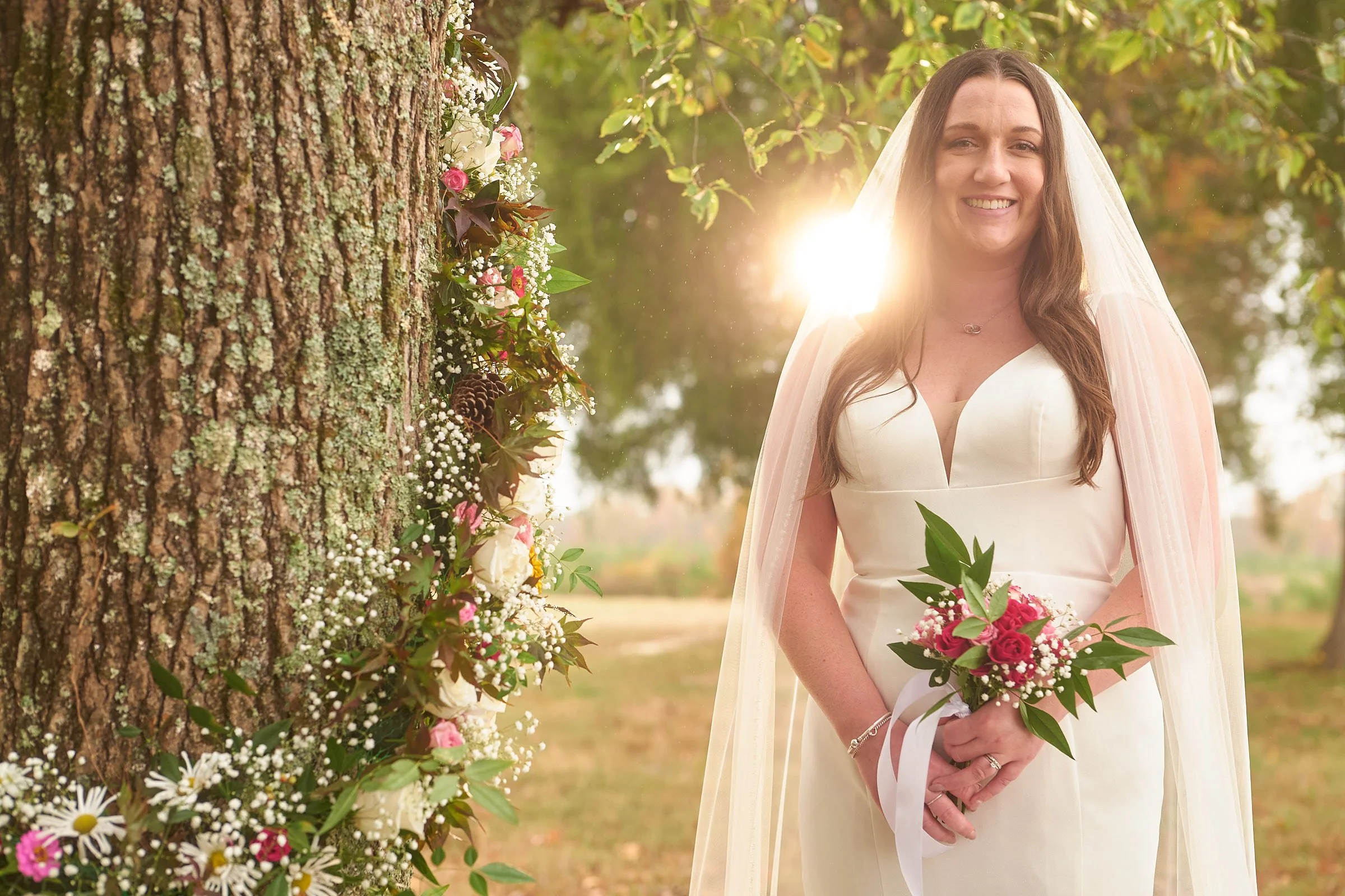 A woman in a wedding dress stands outdoors next to a tree decorated with flowers, smiling as sunlight shines behind her.