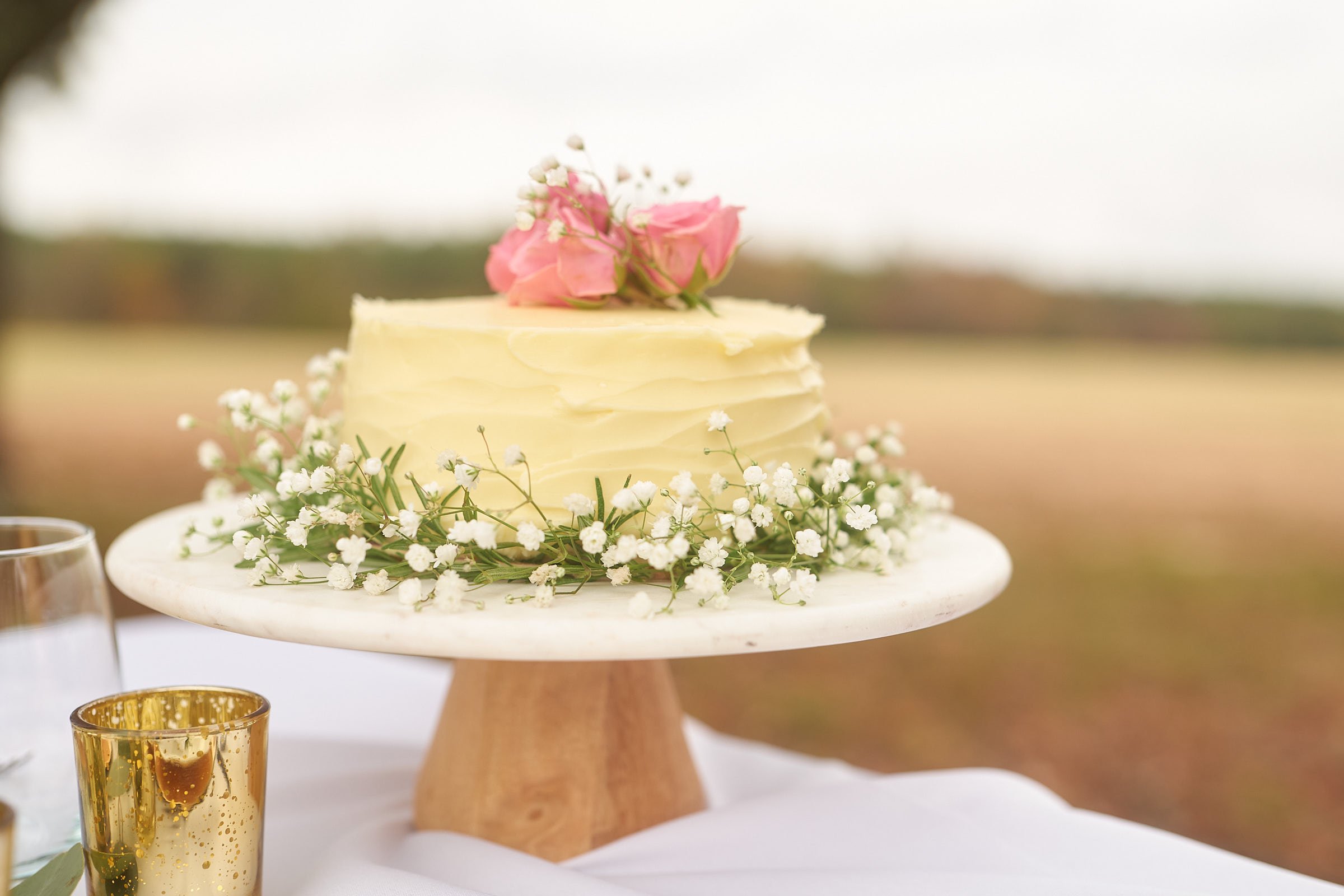 A wedding cake decorated with pink roses and small white flowers, placed on a wooden stand. The cake has a ruffled texture and is set outdoors on a table.