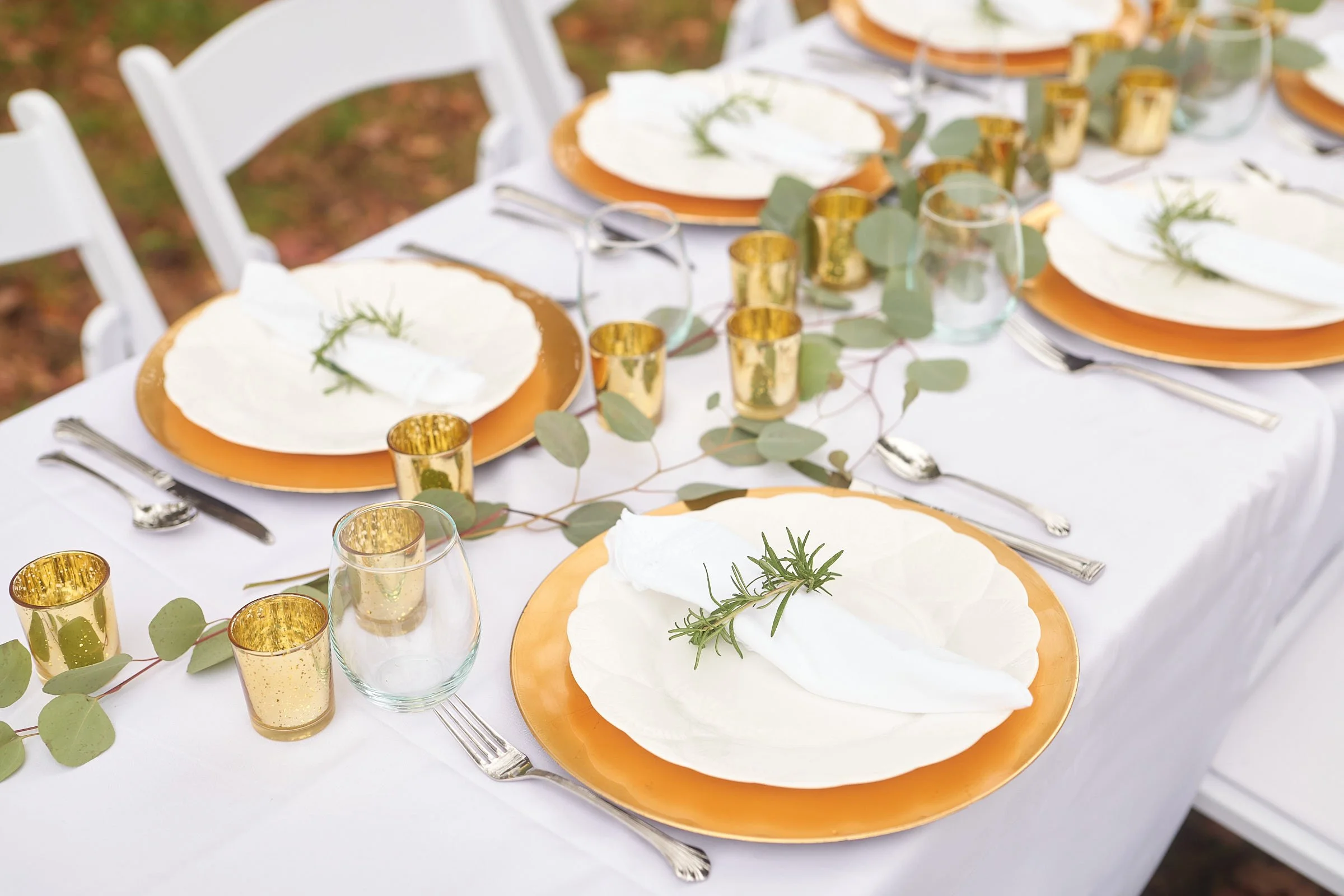 Elegant outdoor table setting with white plates, gold-rimmed glasses, greenery, and a white tablecloth.
