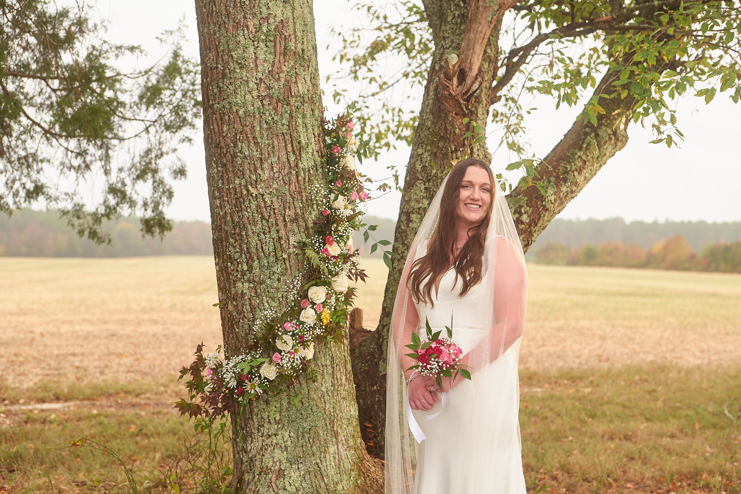 A smiling bride in a white dress with a veil holding a bouquet of pink and white flowers, standing outdoors beside a tree decorated with a floral arrangement.