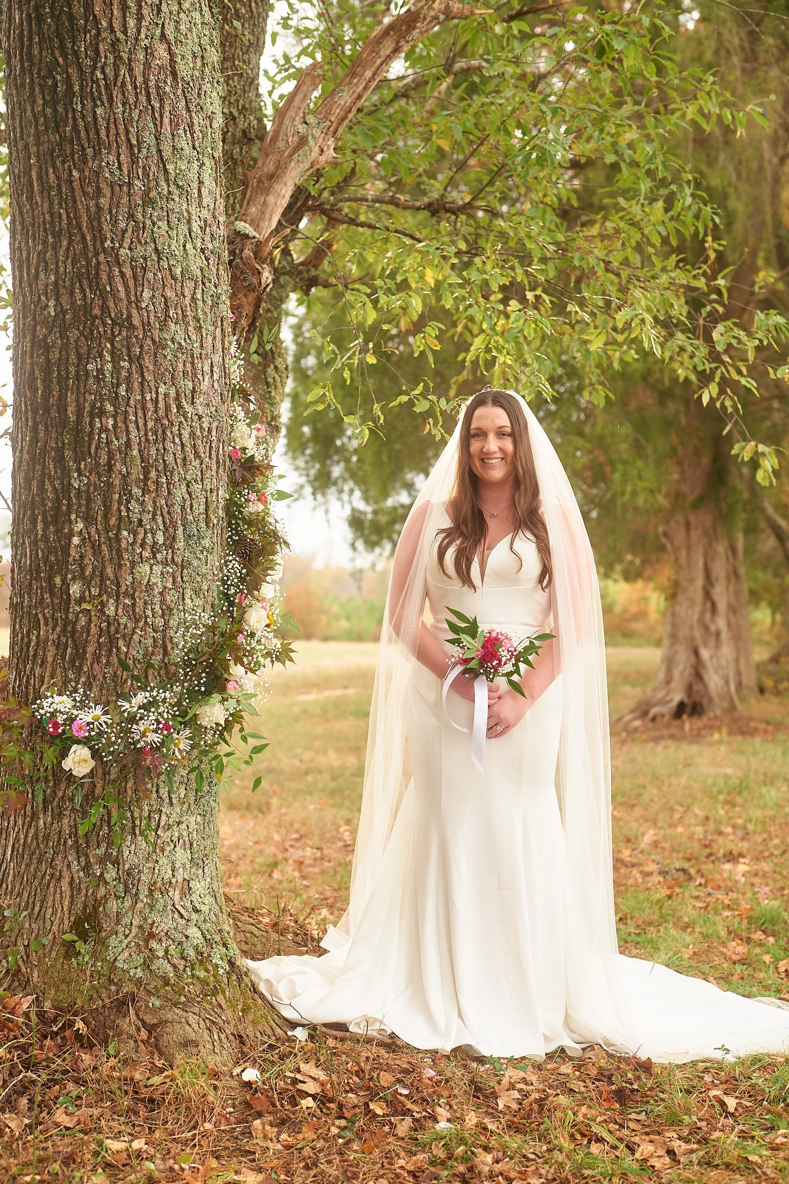 A bride in a white wedding dress and veil holding a bouquet of pink and white flowers, standing outdoors next to a large tree decorated with flowers and greenery.
