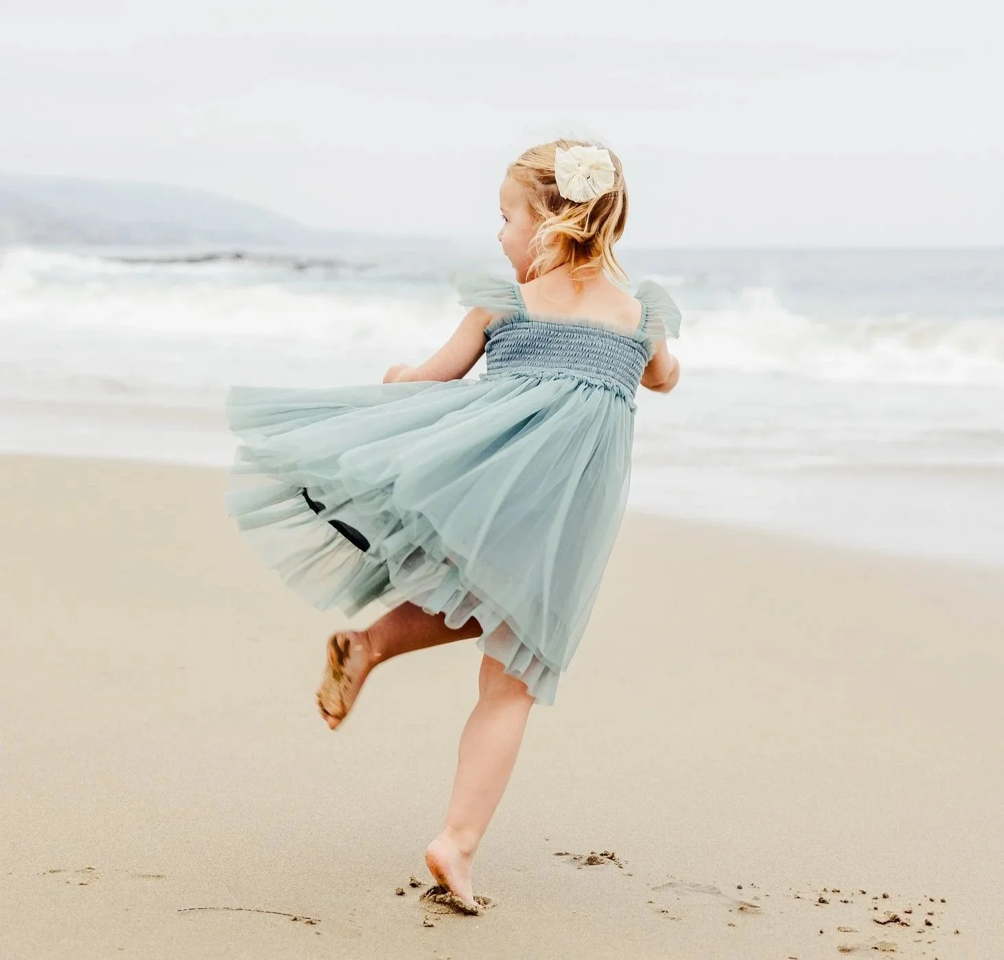 A young girl wearing a light blue dress running barefoot on the beach, with waves in the background.