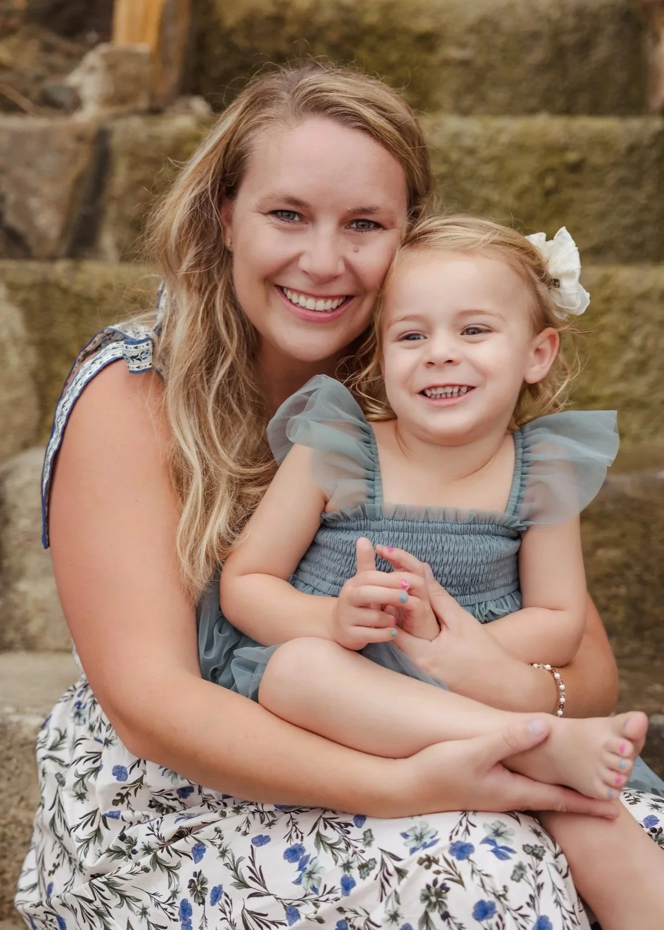 A woman with long blonde hair and a young girl with blonde hair and a white bow in her hair, both smiling, sitting outdoors in front of a stone wall, holding each other.
