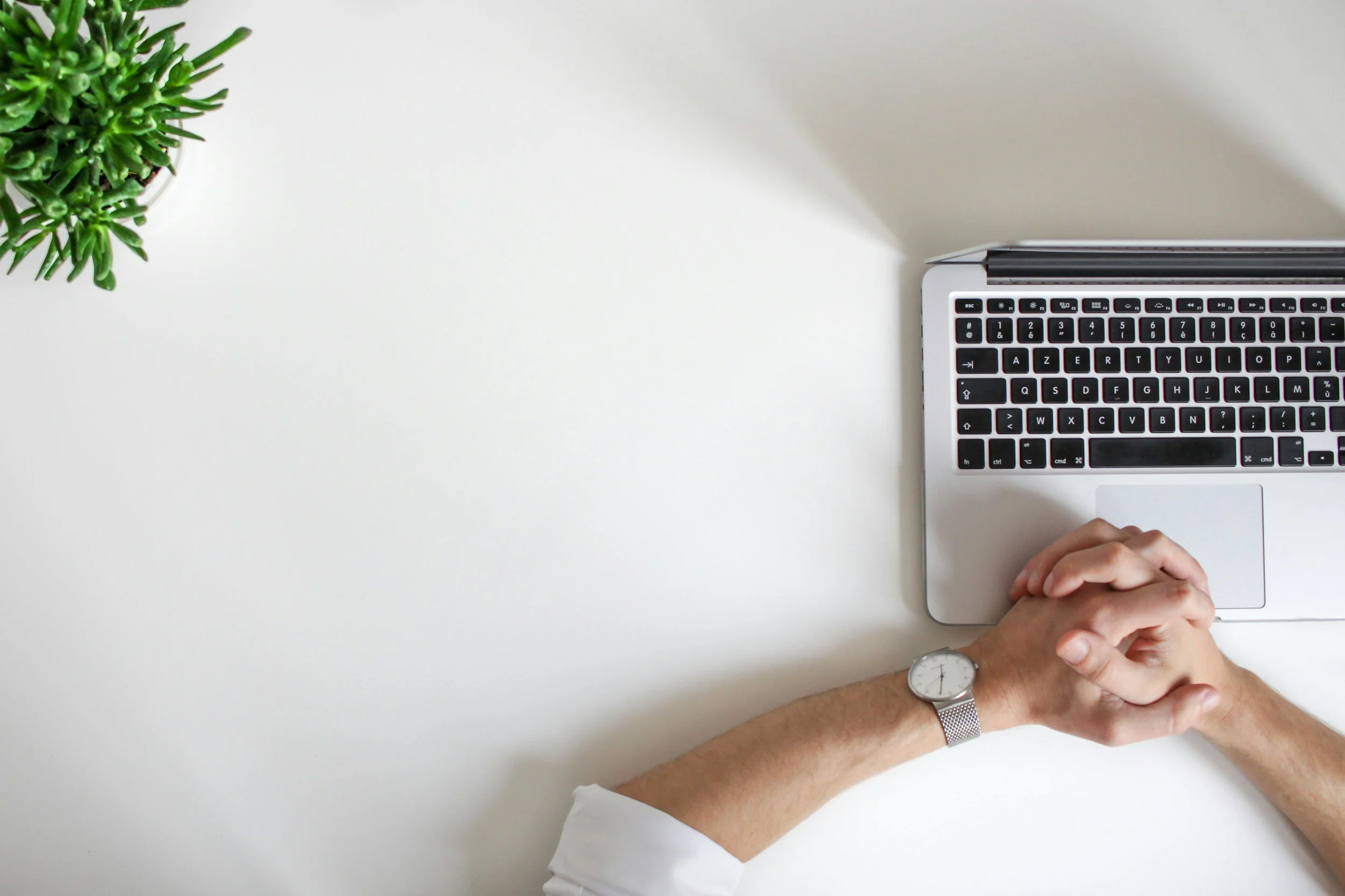 A person's hands with a wristwatch rest on a white desk, next to a silver laptop with black keys, and a green potted plant is partially visible in the top left corner.