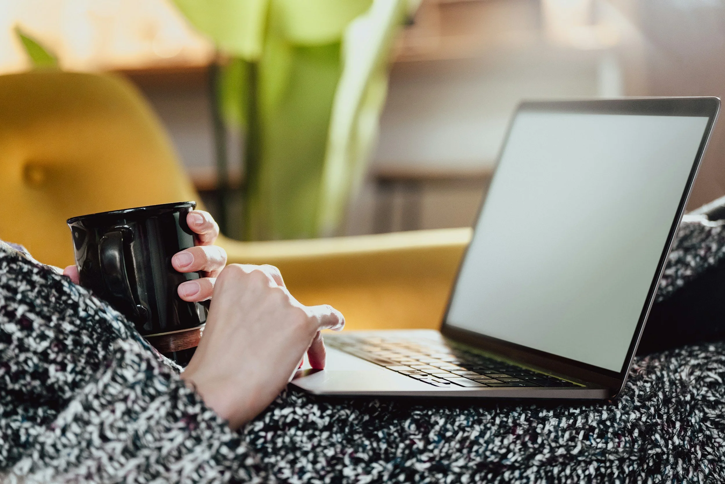 A person holding a black coffee mug while working on a laptop, with a plant in the background.