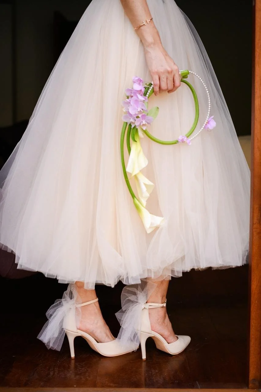 Muñeca vestida con vestido de novia de tul, usando tacones blancos con lazos de tul en los tobillos, sosteniendo un ramo de flores y un collar decorativo.