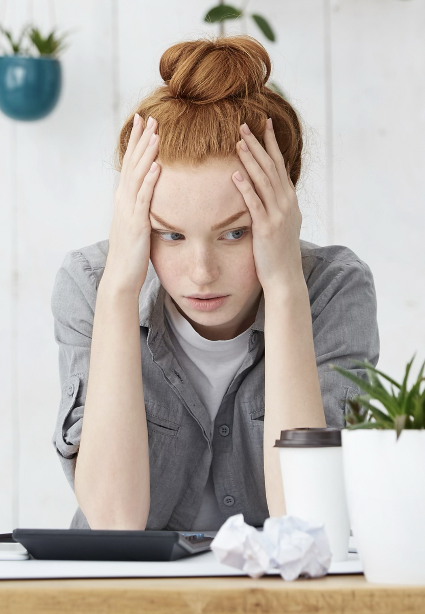 A young woman with red hair and freckles is sitting at a desk, holding her head with both hands, looking stressed or overwhelmed. There are crumpled paper, a disposable coffee cup, and a calculator in front of her, with a plant and a hanging pot in the background.