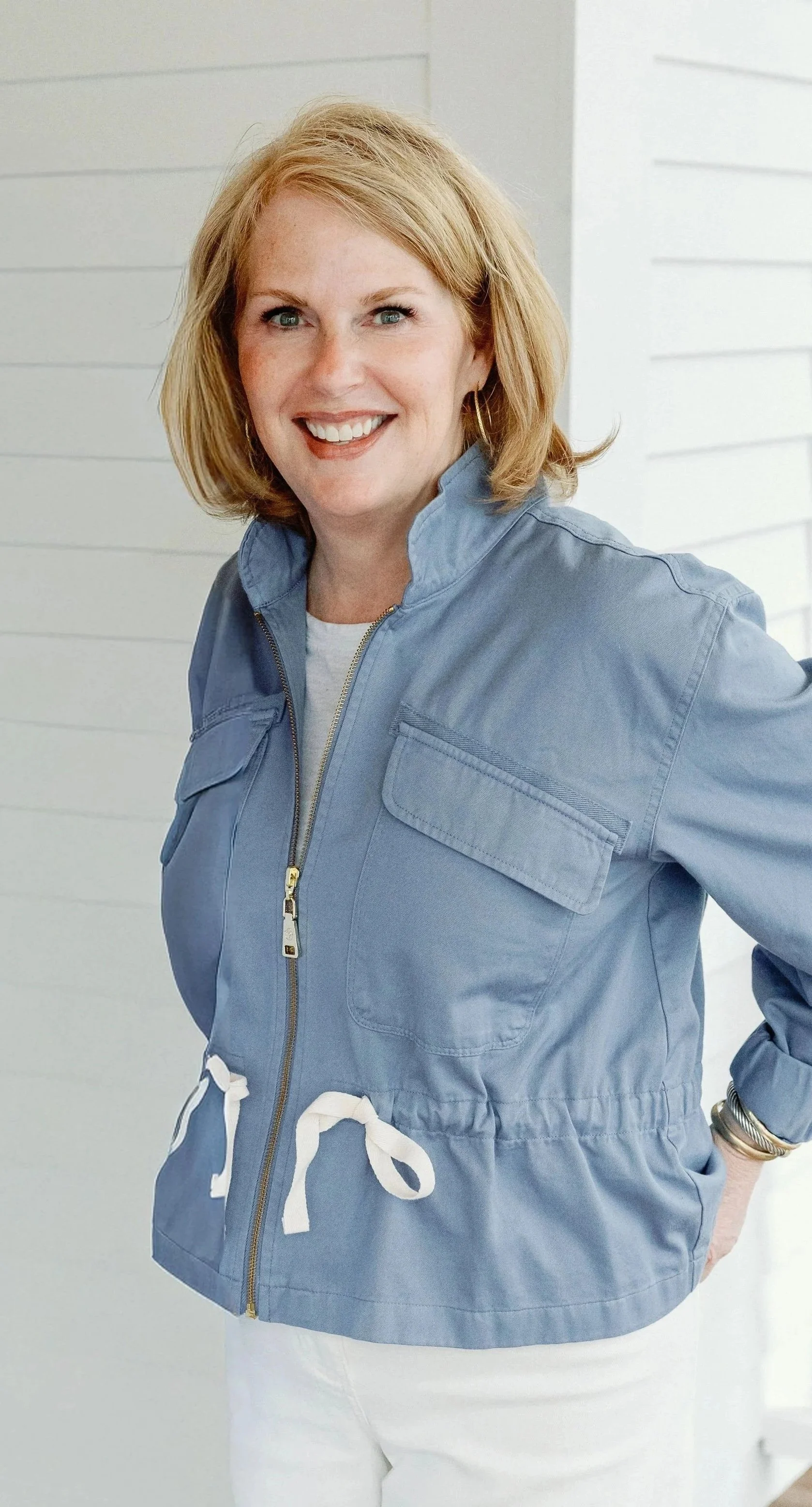 A woman with blonde hair smiling, wearing a blue jacket with a zip and drawstring, standing against a light-colored wall.