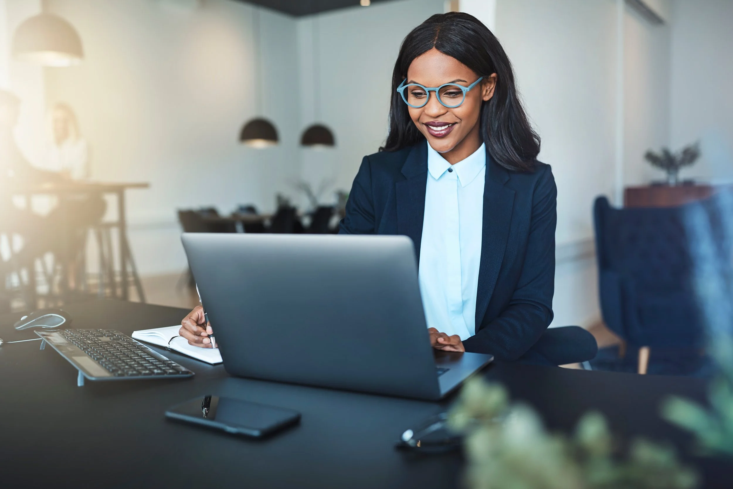 A professional woman with black hair and glasses working at her desk in an office, looking at her laptop screen with a smile.